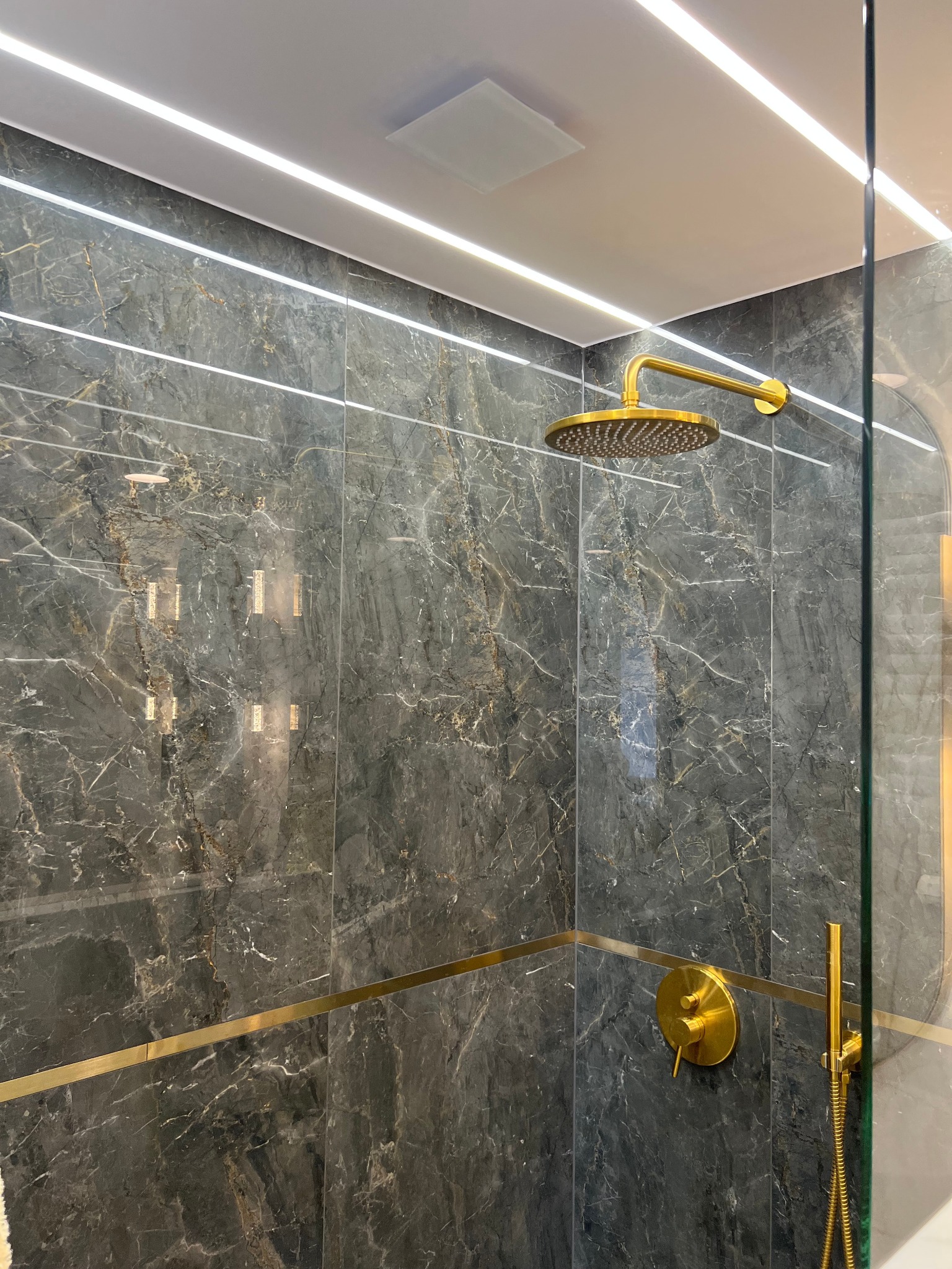Luksusowy prysznic z deszczownicą w kolorze złotym, marmurowe płytki w odcieniach szarości z złotymi akcentami, oświetlenie LED ukryte w suficie, szklana kabina.
