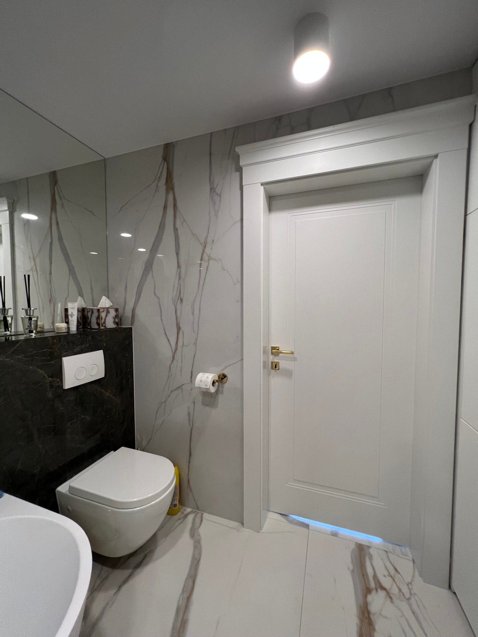 Nowoczesna łazienka z białymi marmurowymi płytkami, podwieszaną toaletą i drzwiami z ozdobną ramą oraz złotymi detalami.