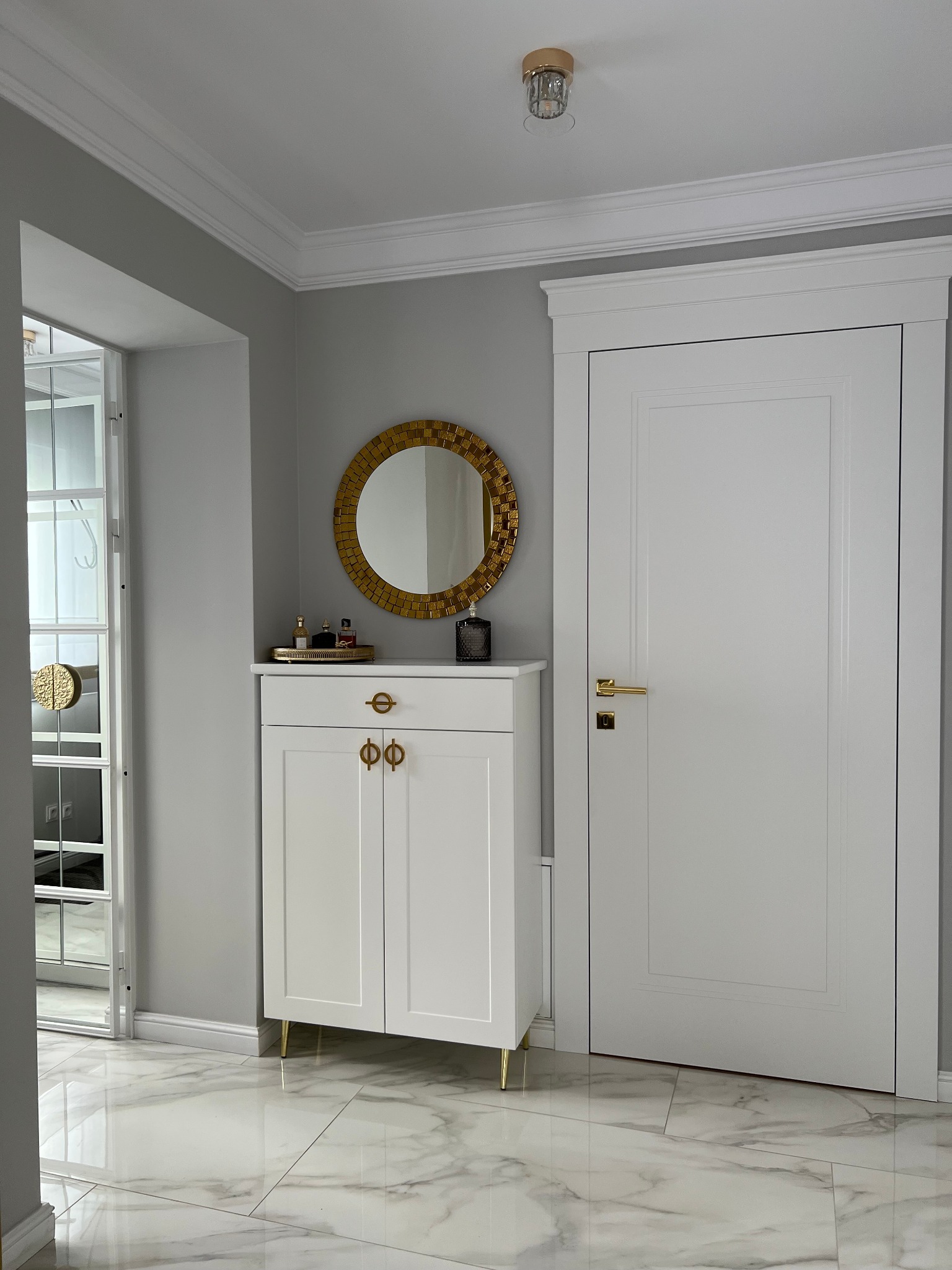 Elegancki przedpokój w odcieniach bieli i szarości z marmurową podłogą, ozdobiony złotymi akcentami na szafce, lustrze i klamkach.