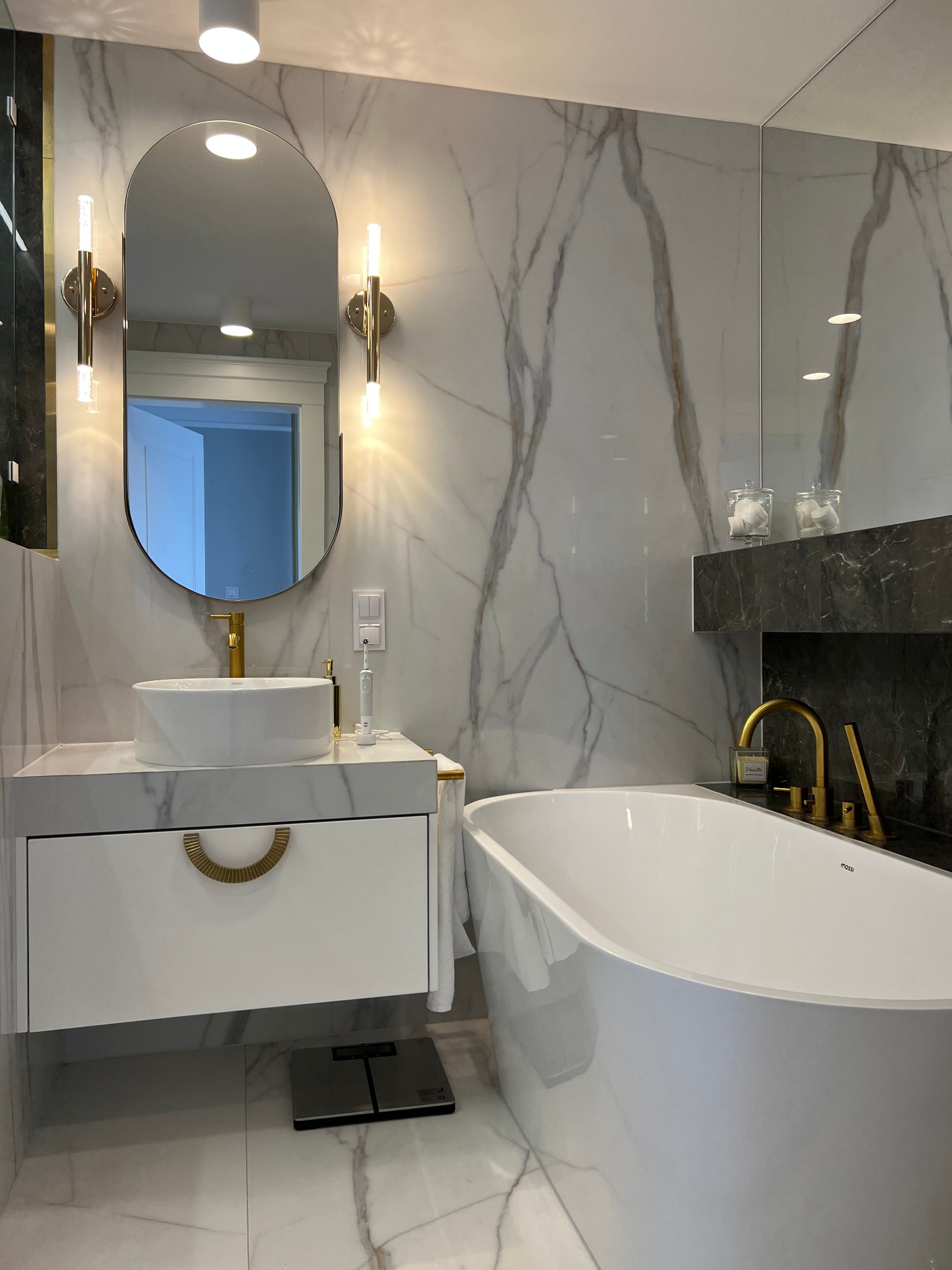 Nowoczesna łazienka z marmurowymi płytkami, owalną umywalką nablatową z baterią w kolorze złota, lustrem z podświetleniem LED i wolnostojącą wanną.