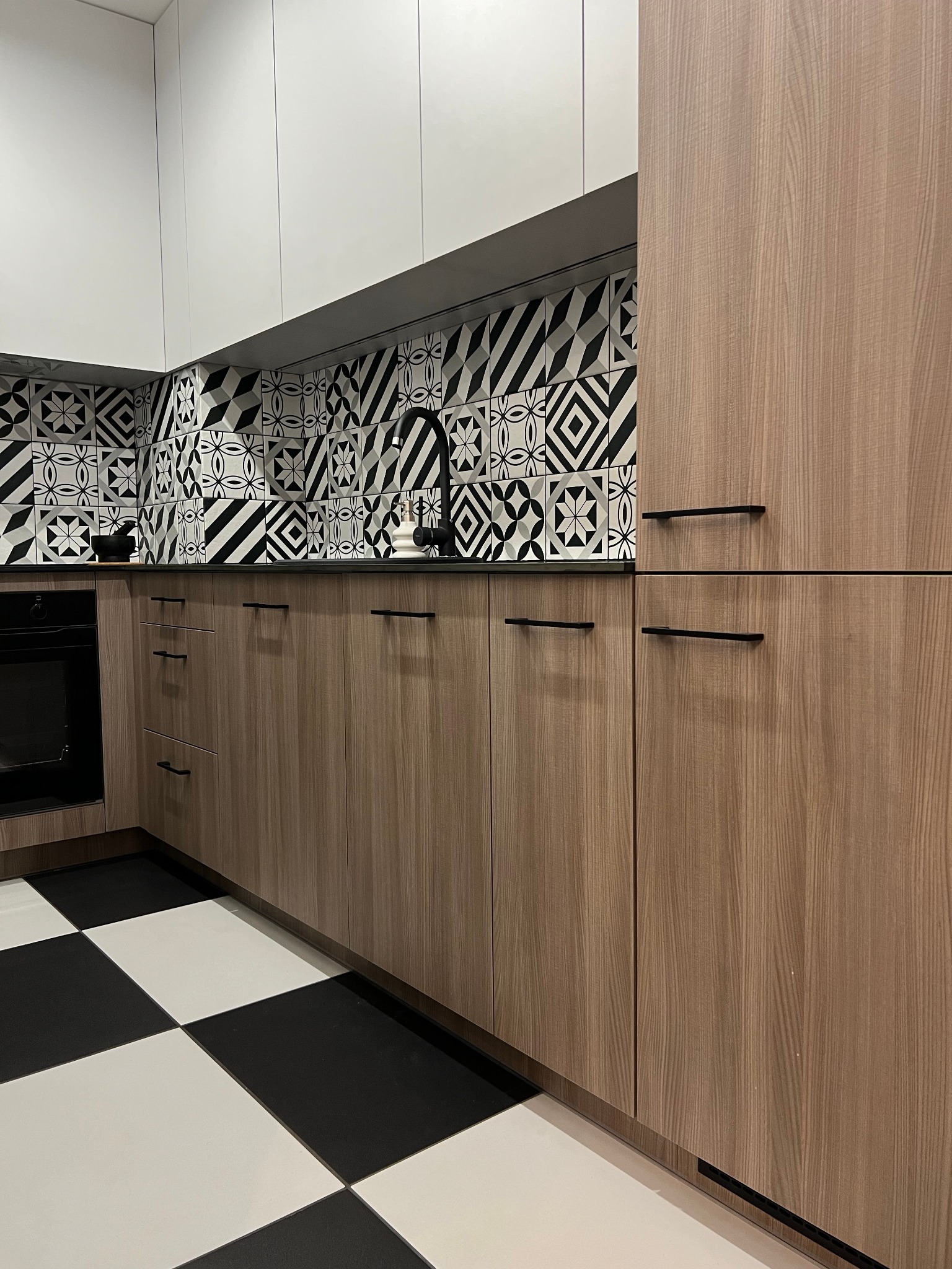 Nowoczesna kuchnia z drewnianymi szafkami, czarnym blatem i mozaiką w czarno-białe wzory na ścianie oraz podłogą w szachownicę.