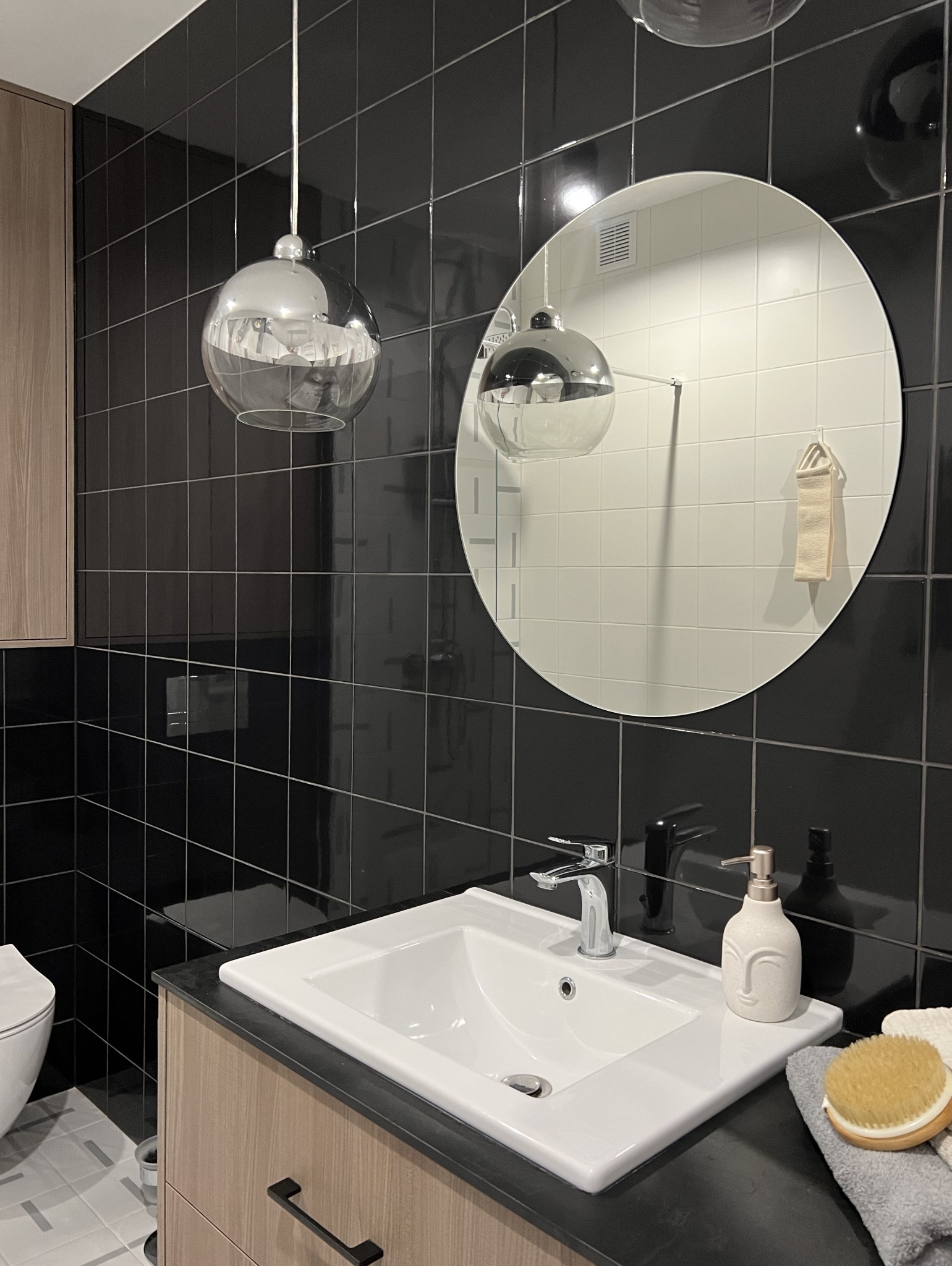 Nowoczesna łazienka z czarnymi płytkami, okrągłym lustrem odbijającym białe płytki i designerskimi lampami wiszącymi w kształcie kuli nad białą umywalką z czarnym blatem i drewnopodobną szafką.