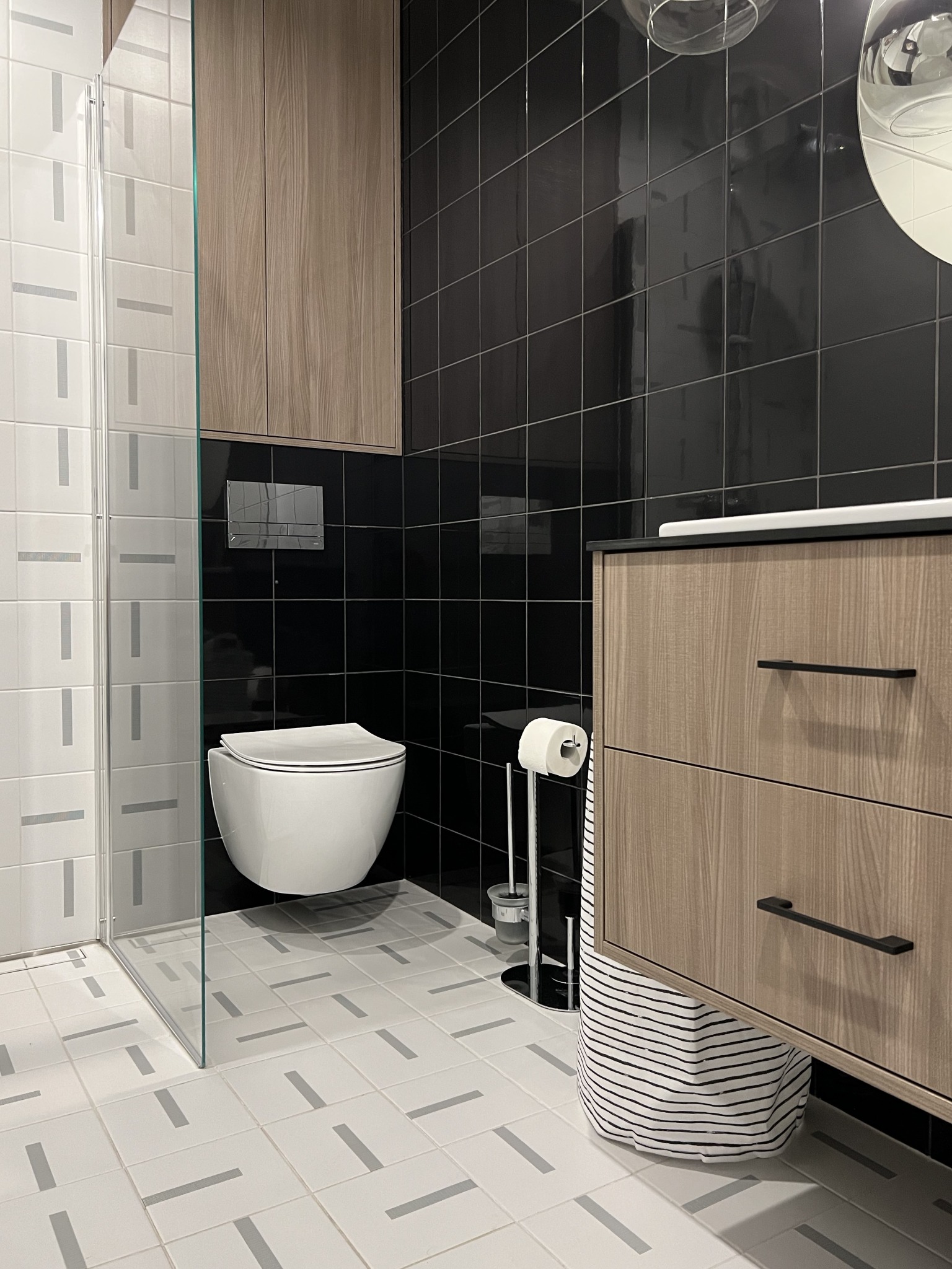Nowoczesna łazienka z czarno-białymi płytkami, podwieszaną toaletą i szafką z drewnopodobnymi frontami oraz koszem na pranie w paski.
