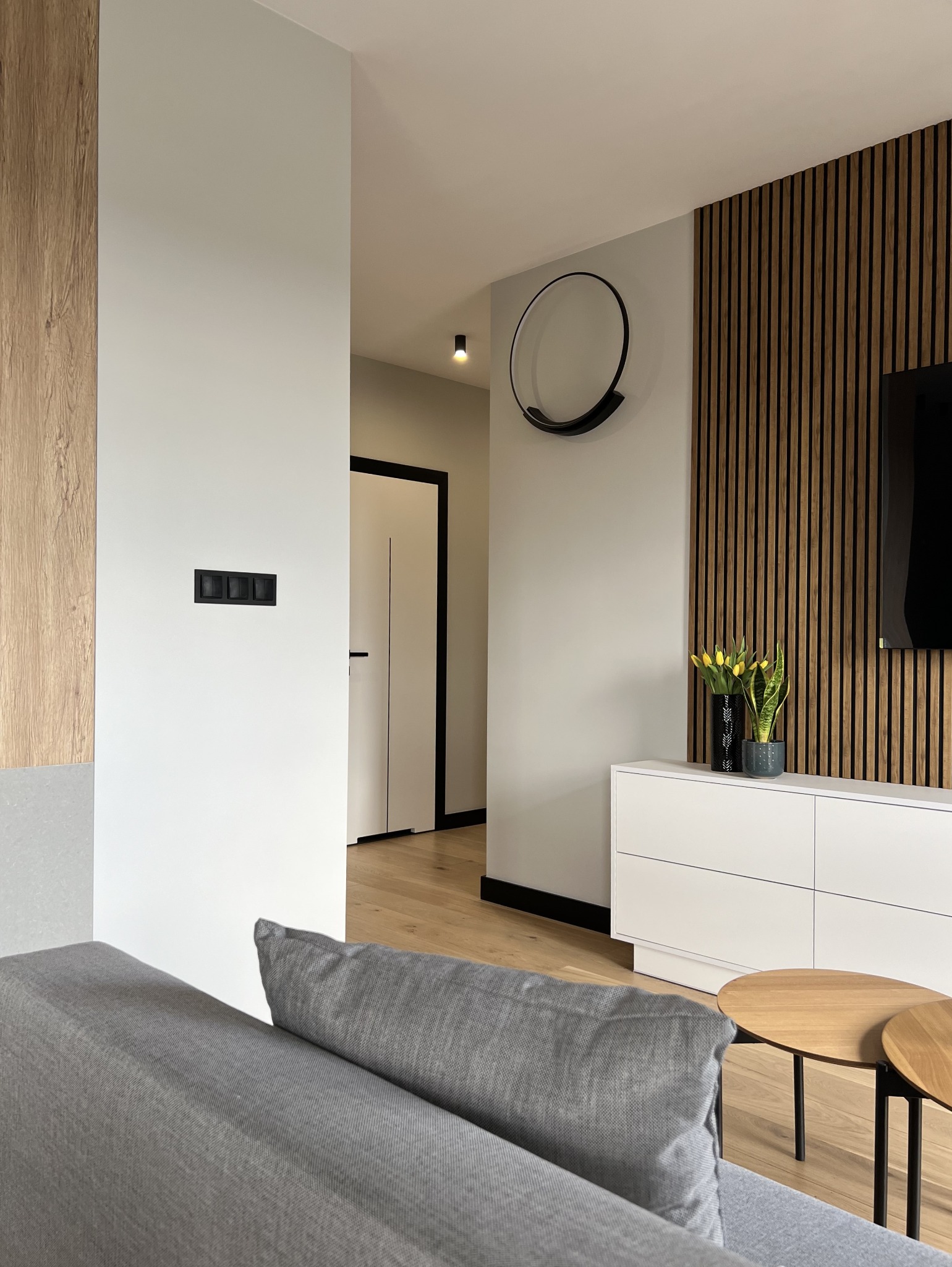 Nowoczesne wnętrze: szara sofa, drewniane lamele na ścianie, biała komoda z dekoracjami roślinnymi i okrągłe stoliki kawowe, widok na przedpokój z białymi drzwiami.