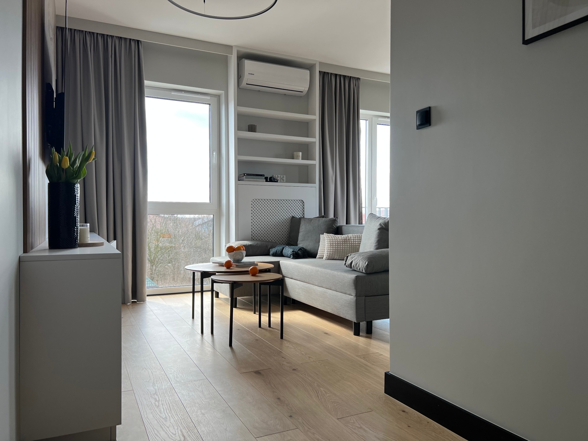 Jasne, nowocześnie urządzone wnętrze salonu z drewnianą podłogą, szarą sofą, okrągłymi stolikami kawowymi i wbudowanym regałem z klimatyzacją.