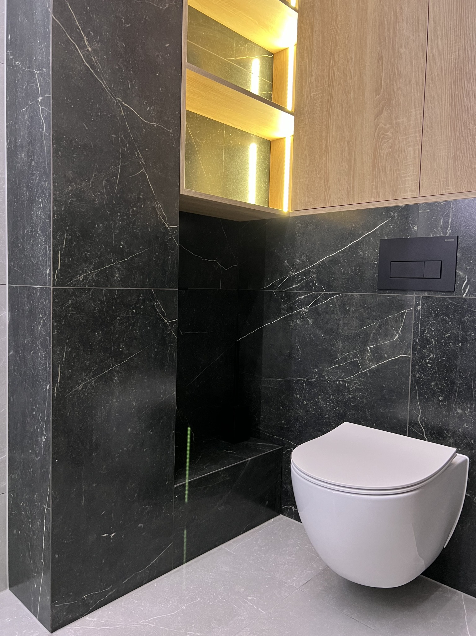 Nowoczesna toaleta podwieszana z czarnymi płytkami imitującymi marmur i drewnianymi szafkami z podświetleniem LED.