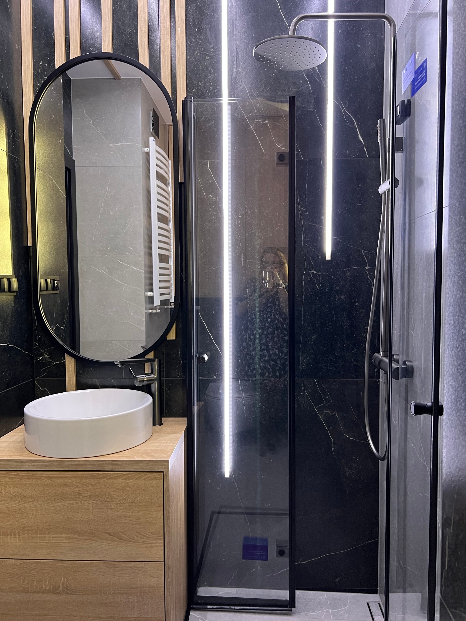 Nowoczesna łazienka z czarnymi marmurowymi płytkami, kabiną prysznicową z podświetleniem LED, owalnym lustrem i drewnianą szafką pod umywalką.