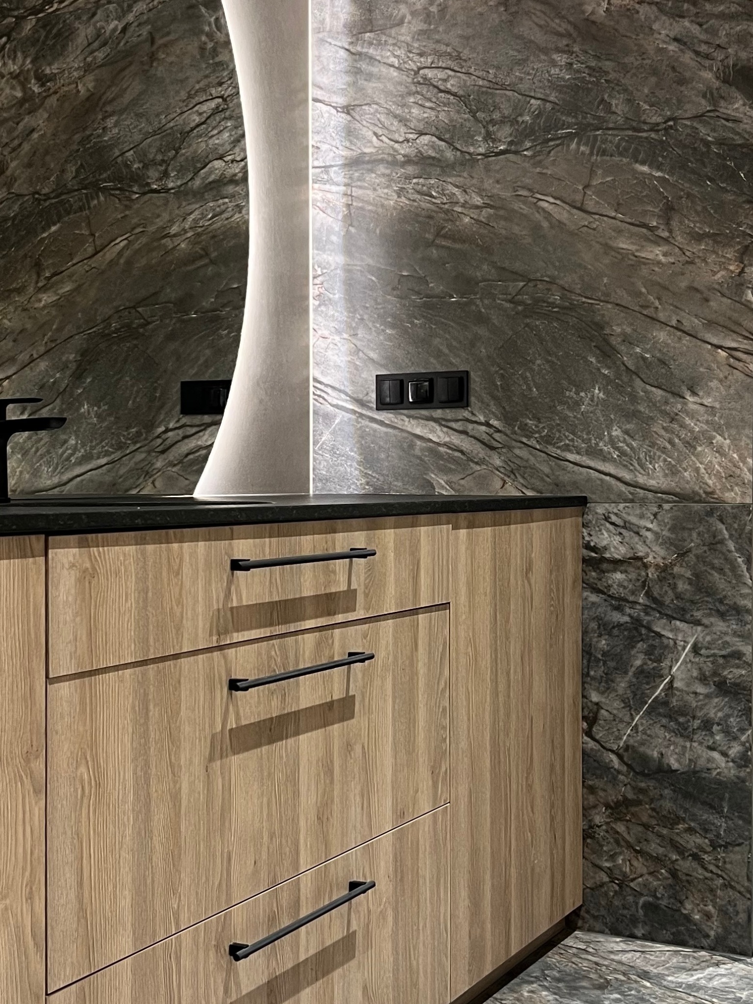 Nowoczesna łazienka z drewnianą szafką pod umywalkę z czarnymi uchwytami, czarnym blatem i marmurową ścianą z podświetleniem LED.