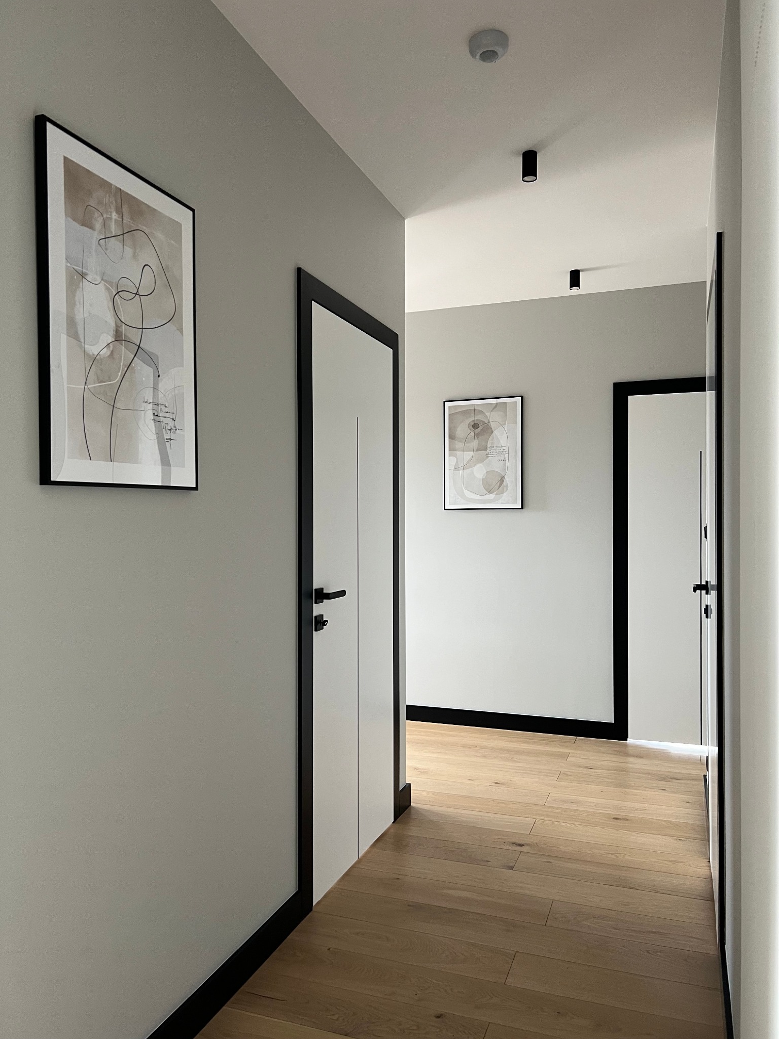 Korytarz w Tomaszowie Mazowieckim po remoncie, szare ściany, drewniana podłoga, białe drzwi z czarnymi ościeżnicami, abstrakcyjne obrazy na ścianach.