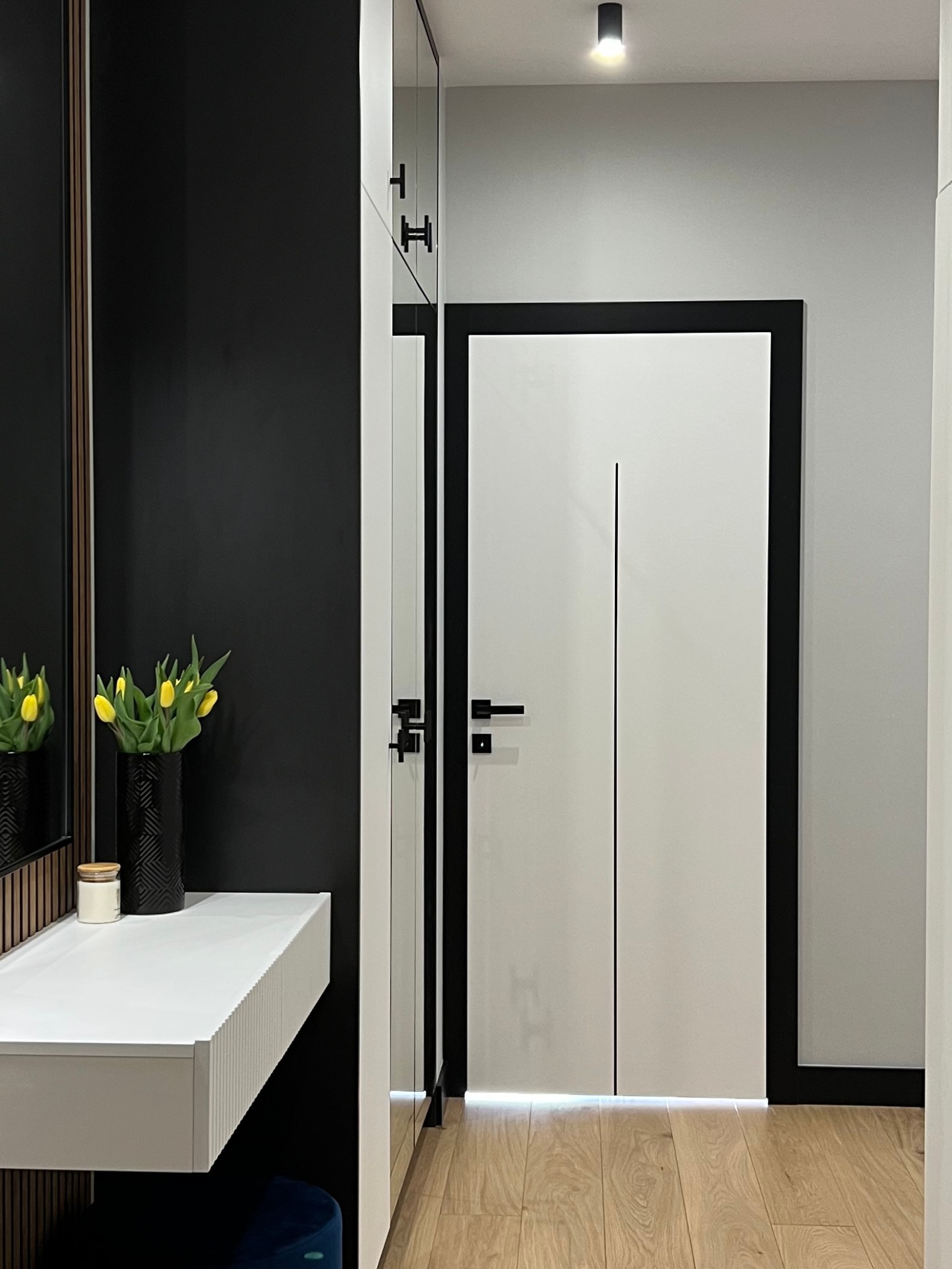 Nowoczesny przedpokój z białymi drzwiami z czarną ramą, lustrem, konsolą z żółtymi tulipanami w czarnym wazonie i drewnianą podłogą.