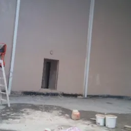 Wnętrze hali z nałożonymi tynkami, widoczne ślady prac na podłodze, drabina, wiaderka i resztki materiałów budowlanych.