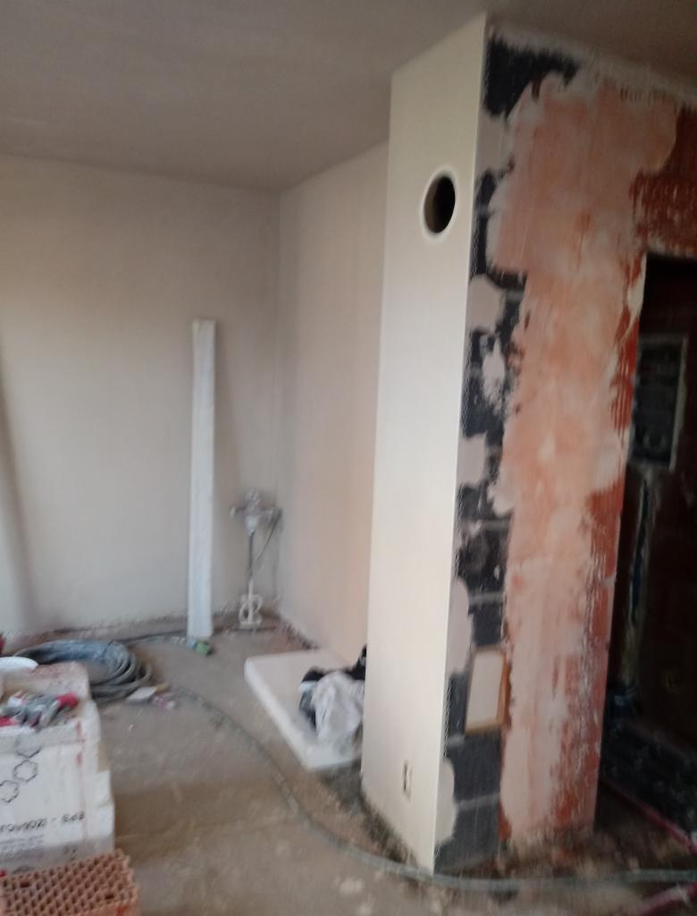 Wnętrze w trakcie remontu: fragment ściany z fragmentami starego tynku, obok pomalowany filar z otworem wentylacyjnym, widoczne narzędzia i materiały budowlane na podłodze.