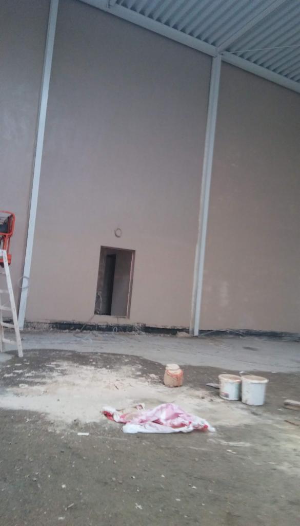 Wnętrze hali z nałożonymi tynkami, widoczne ślady prac na podłodze, drabina, wiaderka i resztki materiałów budowlanych.