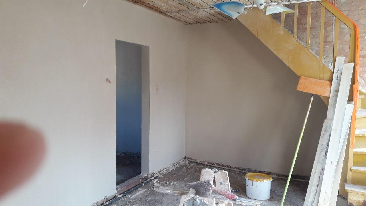 Pokój w trakcie prac wykończeniowych: świeżo nałożone tynki na ścianach, widoczne narzędzia murarskie na podłodze, schody z żółtą barierką i nieotynkowany sufit.