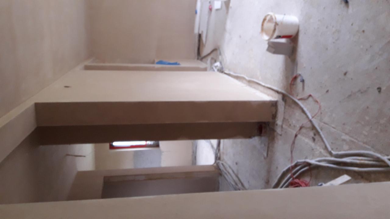 Wnętrze w trakcie prac wykończeniowych: świeżo nałożona, jasna warstwa na ścianach i suficie, widoczne przewody elektryczne, wiaderko na ścianie i surowa podłoga.