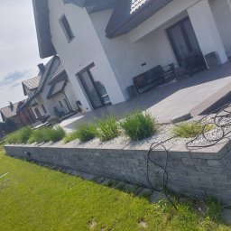 Ad-bruk - Ogród przydomowy z podwyższonym tarasem, rabatą z roślinami ozdobnymi i kamieniami, oraz niskim murkiem z szarej kostki. Widoczny dom z dużym oknem tarasowym.