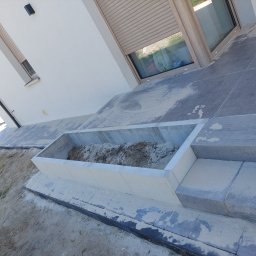 Ad-bruk - Betonowa donica wbudowana w taras z szarej kostki, wypełniona ziemią, widok z góry na tle białej ściany budynku z oknami z roletami.