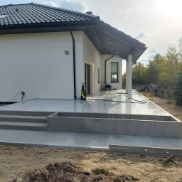 Ad-bruk - Nowoczesny taras z jasnych płyt, schody z obrzeżami w Woli Moszczenickiej, widoczne prace wykończeniowe i sprzęt do czyszczenia.