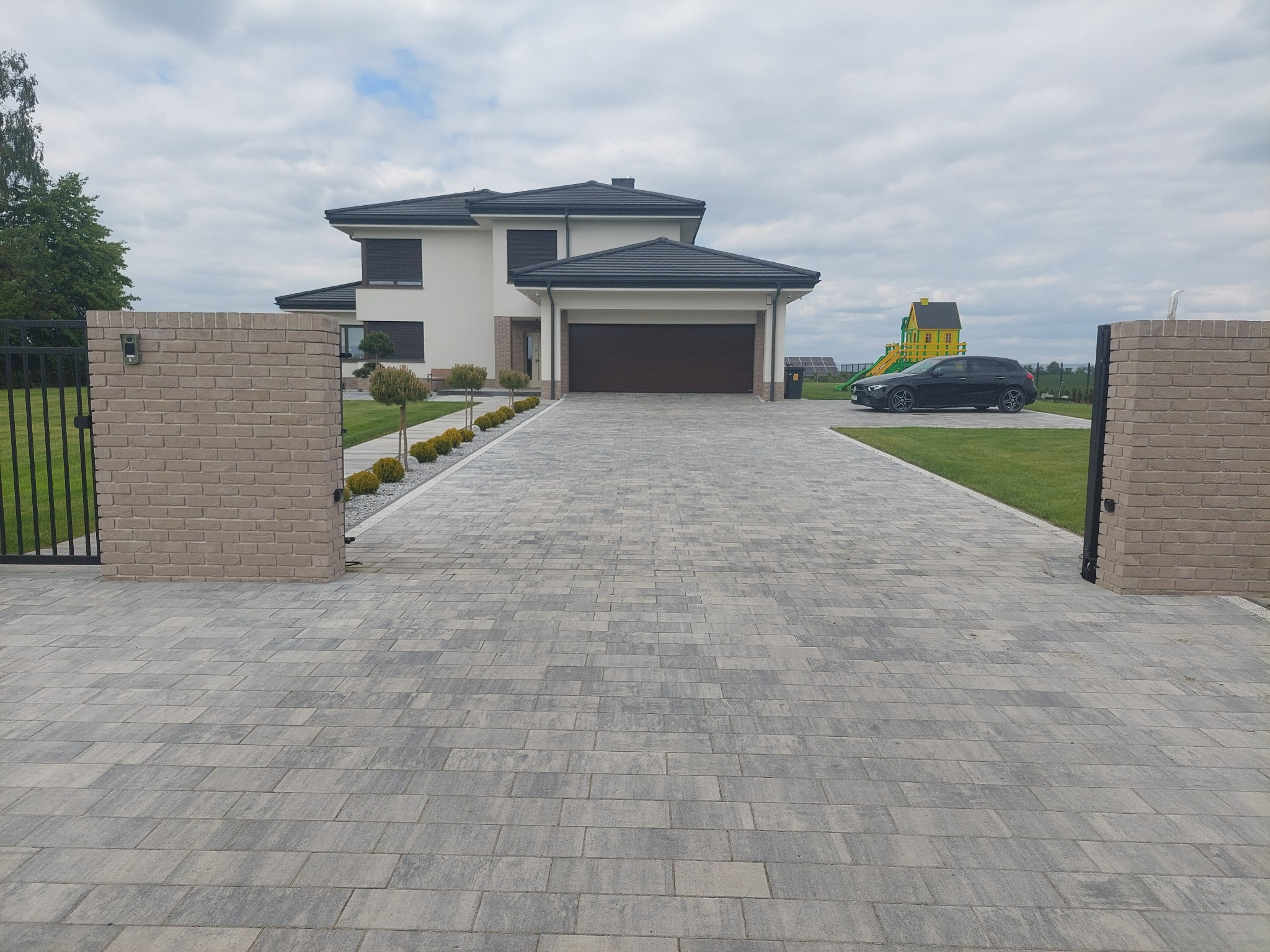 Szeroki kadr na nowoczesny dom z dwustanowiskowym garażem i podjazdem z szarej kostki, otoczony zielonym trawnikiem, z bramą wjazdową z ceglanymi słupkami.