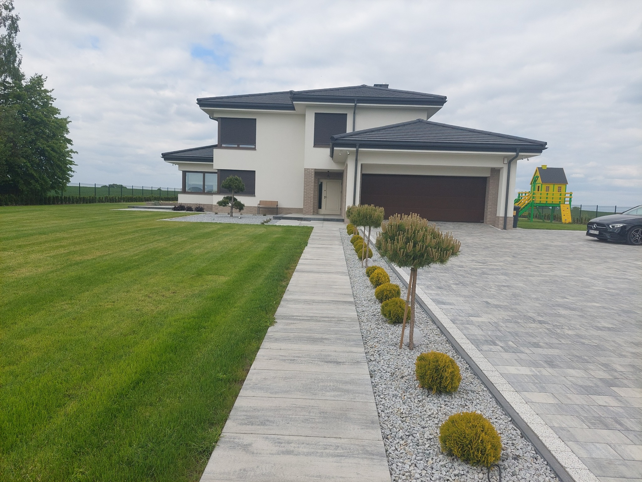 Nowoczesny dom jednorodzinny z brukowanym podjazdem i chodnikiem z płyt, ozdobnymi krzewami i trawnikiem, plac zabaw w tle.