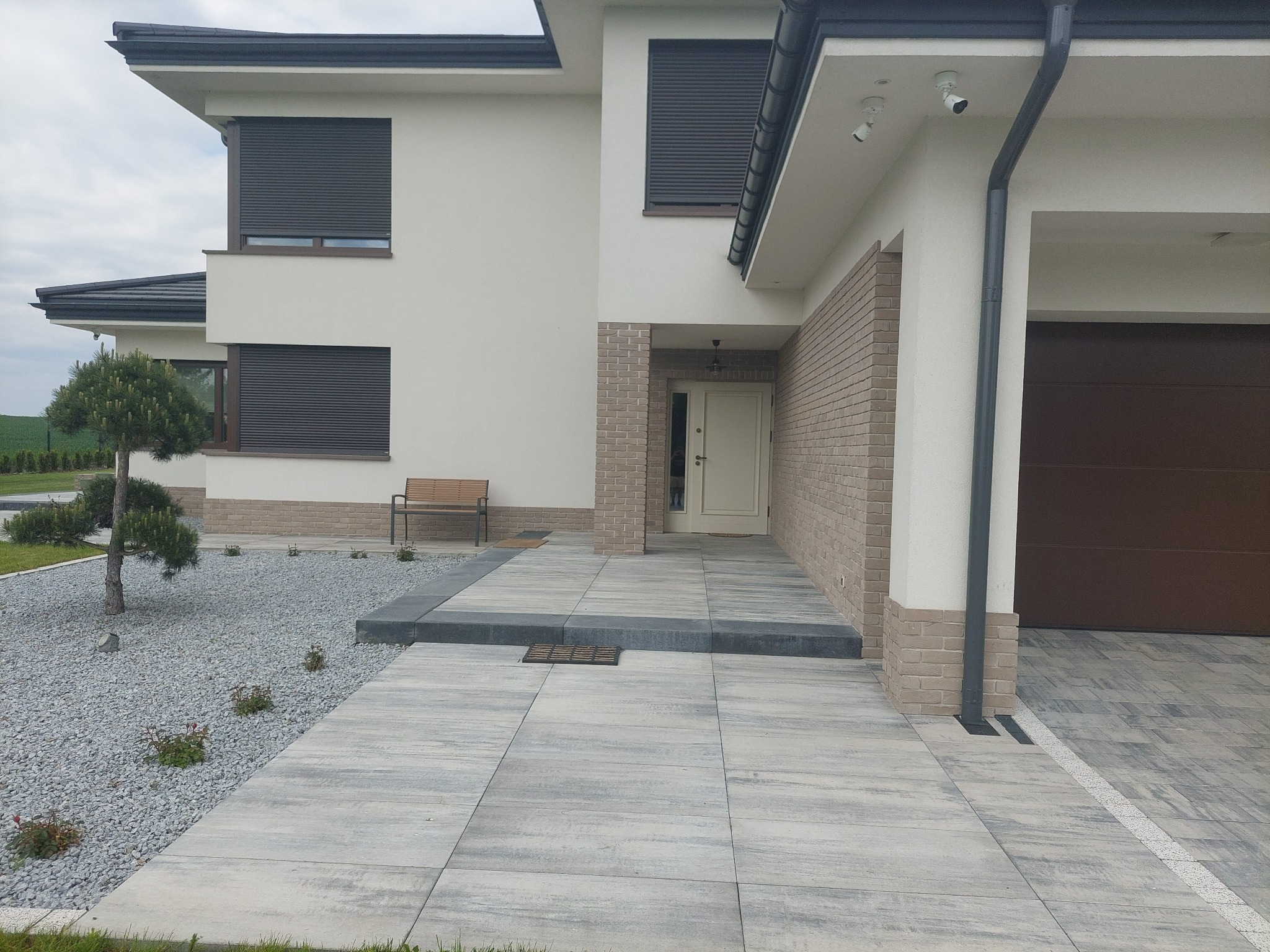 Nowoczesny dom jednorodzinny z podjazdem wyłożonym szarą kostką w dużym formacie, obok ozdobny ogródek z kamykami i bonsai.