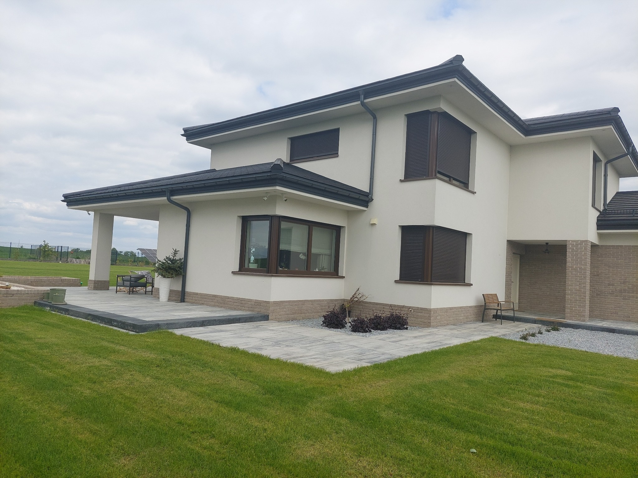 Nowoczesny dom z białymi ścianami i ciemnymi okiennicami, otoczony świeżo ułożoną szarą kostką brukową i zielonym trawnikiem, widok z perspektywy trawnika.