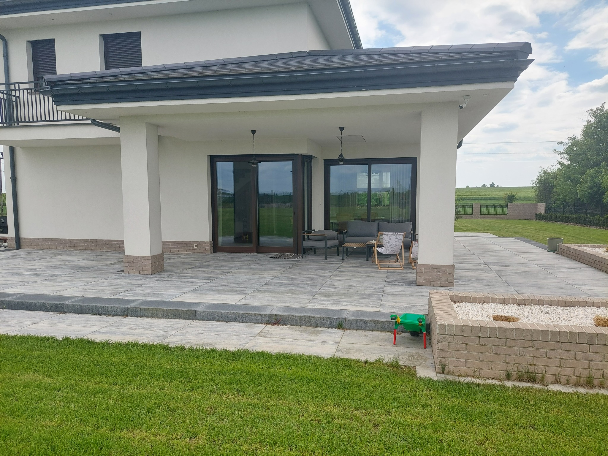 Nowoczesny taras z jasnoszarej kostki brukowej, meble ogrodowe pod zadaszeniem domu, widok na zadbany trawnik i rabaty kwiatowe, zabawka dziecka przy rabacie.