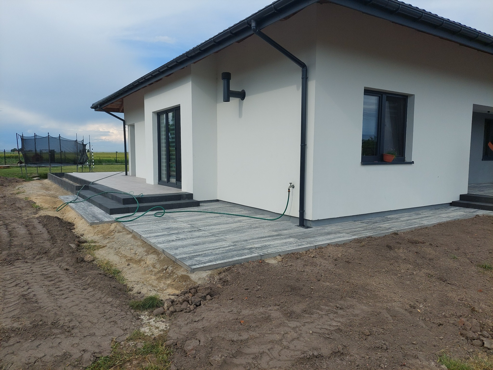 Nowoczesny dom z szarą kostką brukową ułożoną na tarasie i ścieżce, widoczny fragment trawnika z trampoliną w tle oraz czarne rury spustowe na białej elewacji.