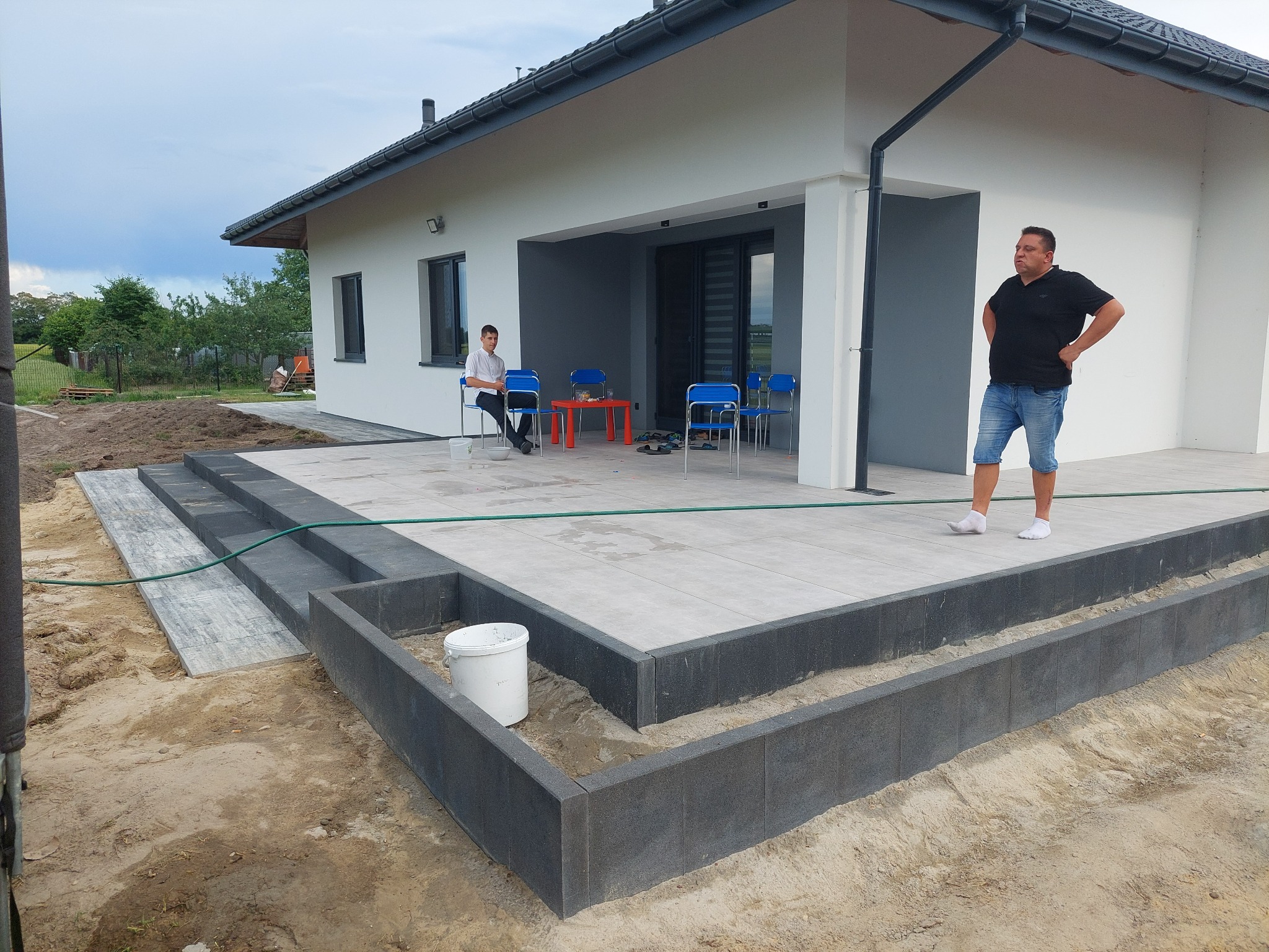 Nowoczesny dom z tarasem wyłożonym jasnymi płytami, widoczne schody z ciemnej kostki, obrzeża rabat z szarej kostki, w tle dwóch mężczyzn.