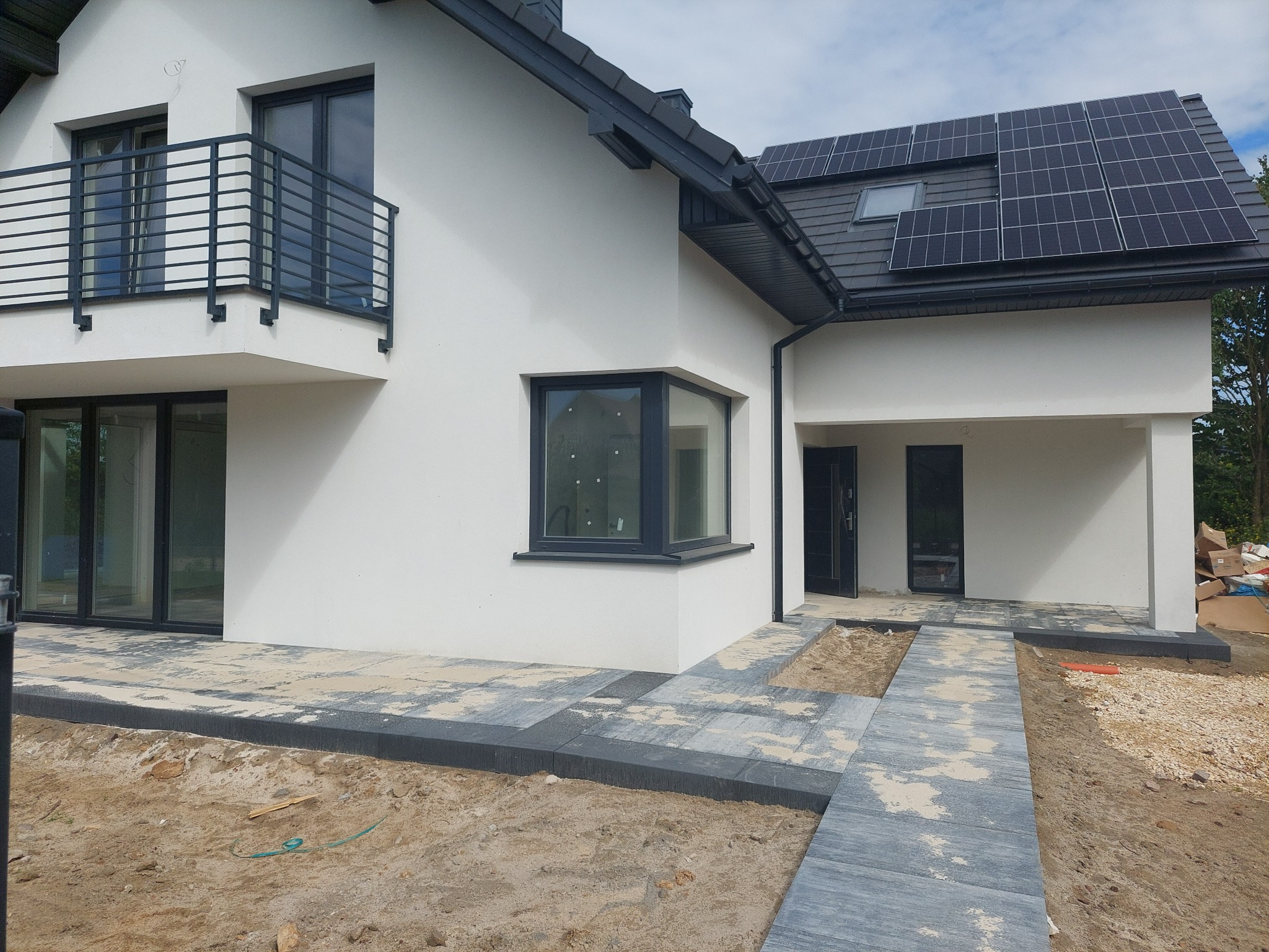 Nowoczesny dom z białymi ścianami, szarą stolarką okienną i panelami słonecznymi na dachu, częściowo wyłożony kostką brukową, z widocznymi śladami piasku budowlanego.