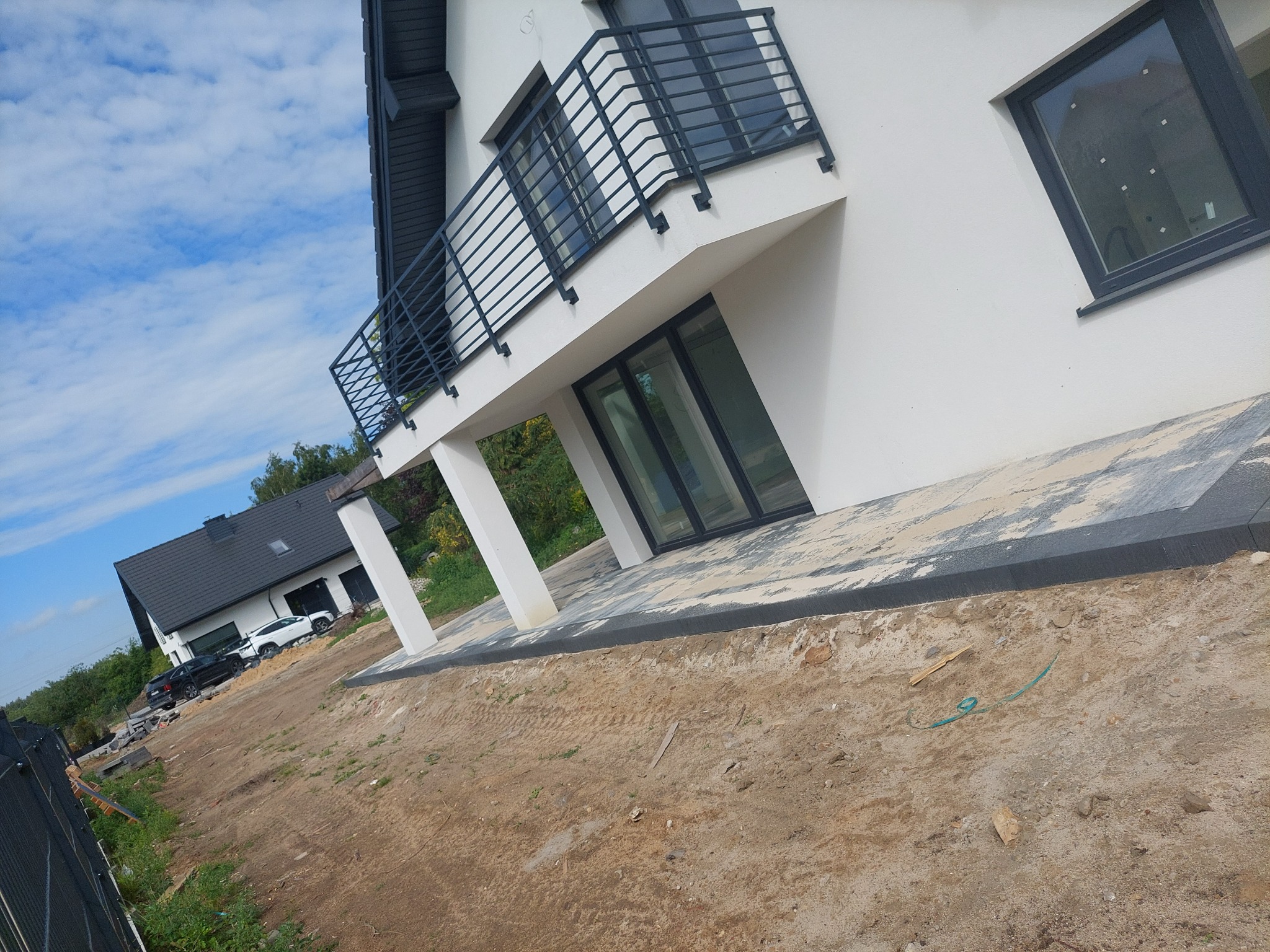 Nowoczesny dom z szarą kostką brukową wokół, widok na balkon z czarną balustradą, w tle drugi budynek i samochody, teren przygotowany pod trawnik.