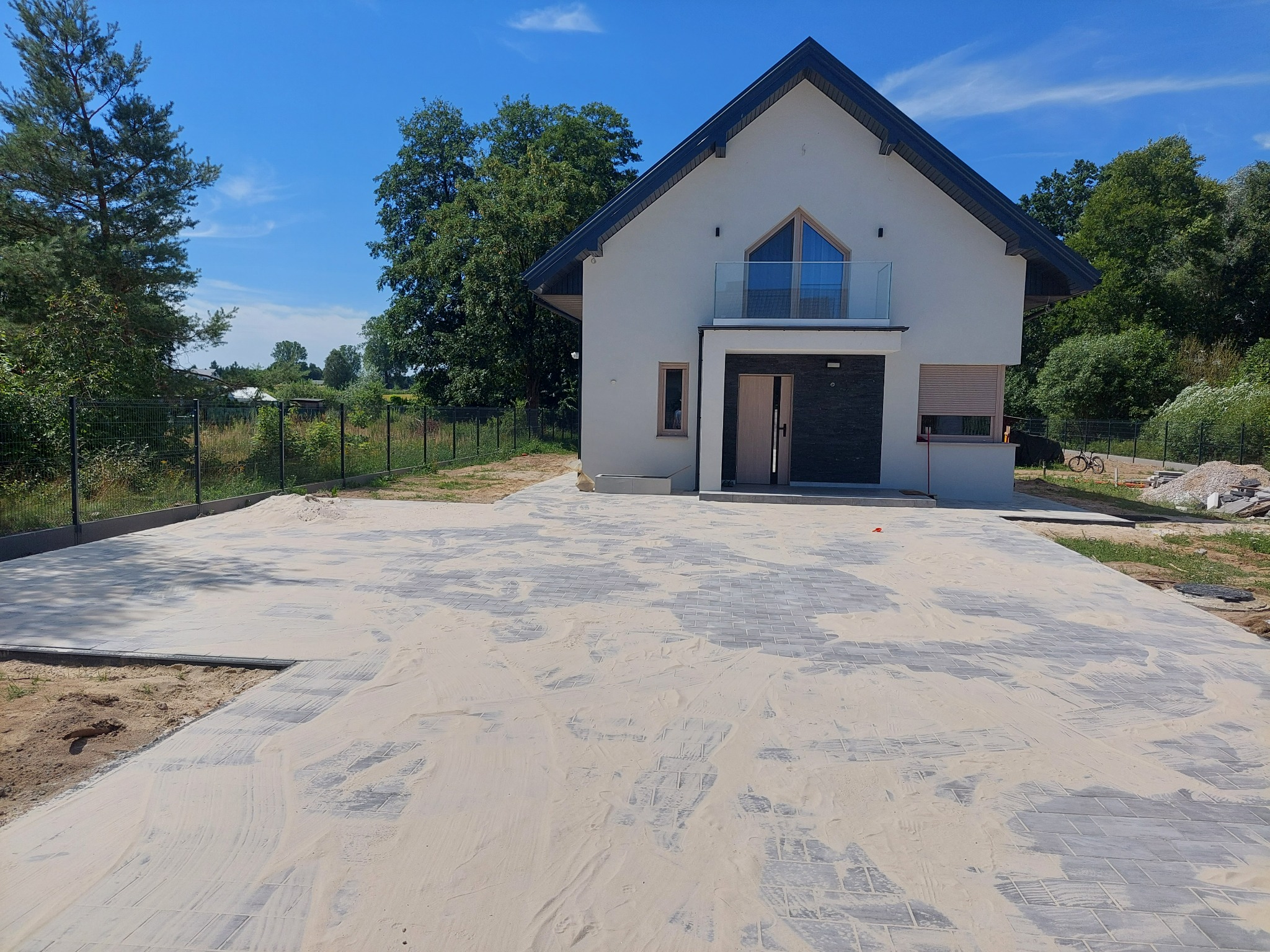 Nowoczesny dom jednorodzinny z szarą kostką brukową na podjeździe, zasypaną piaskiem w trakcie prac wykończeniowych, widok z przodu w słoneczny dzień.