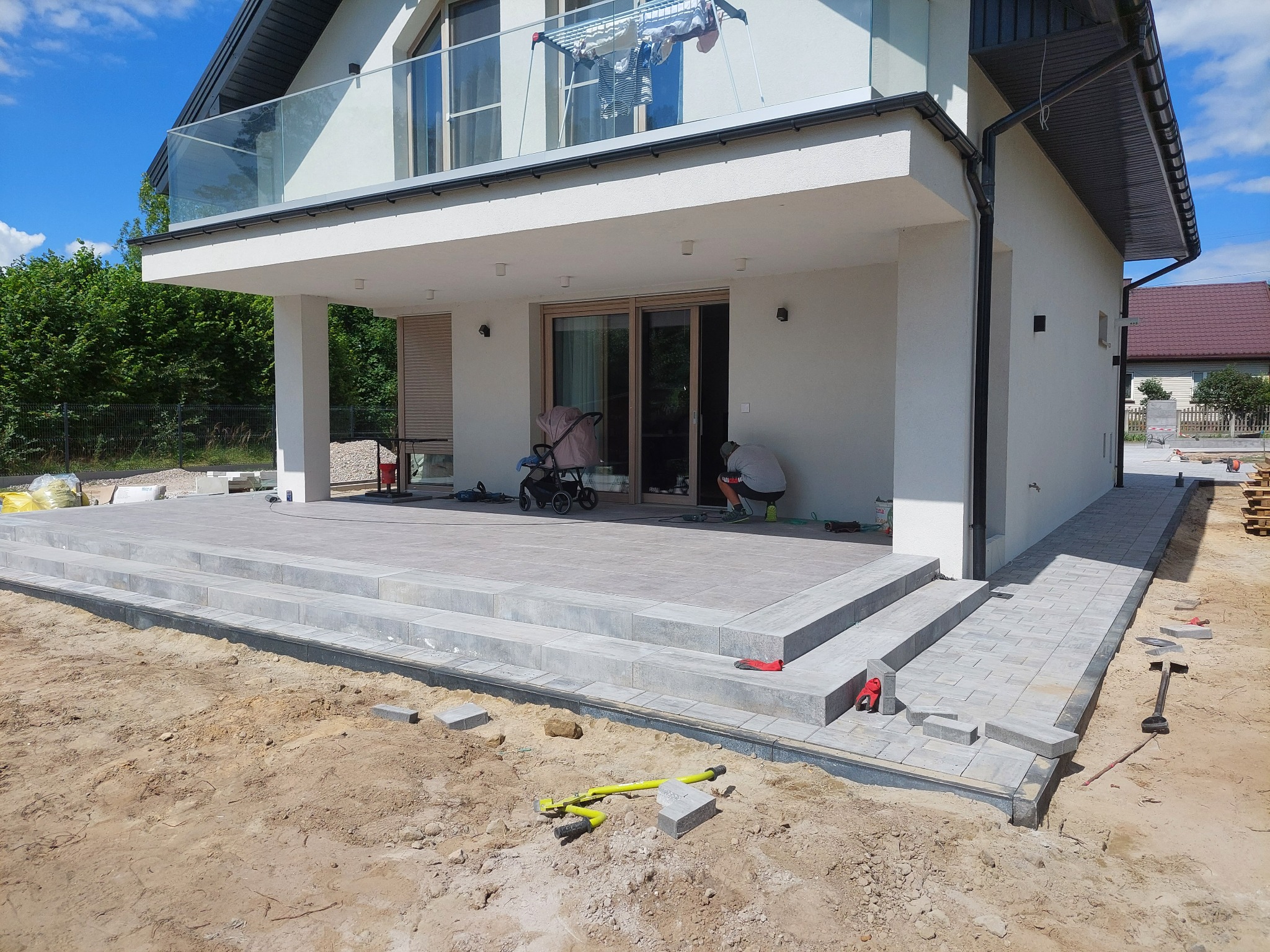 Budowa tarasu z kostki brukowej przed nowoczesnym domem z balkonem, widoczne narzędzia i częściowo ułożona nawierzchnia, praca w toku.