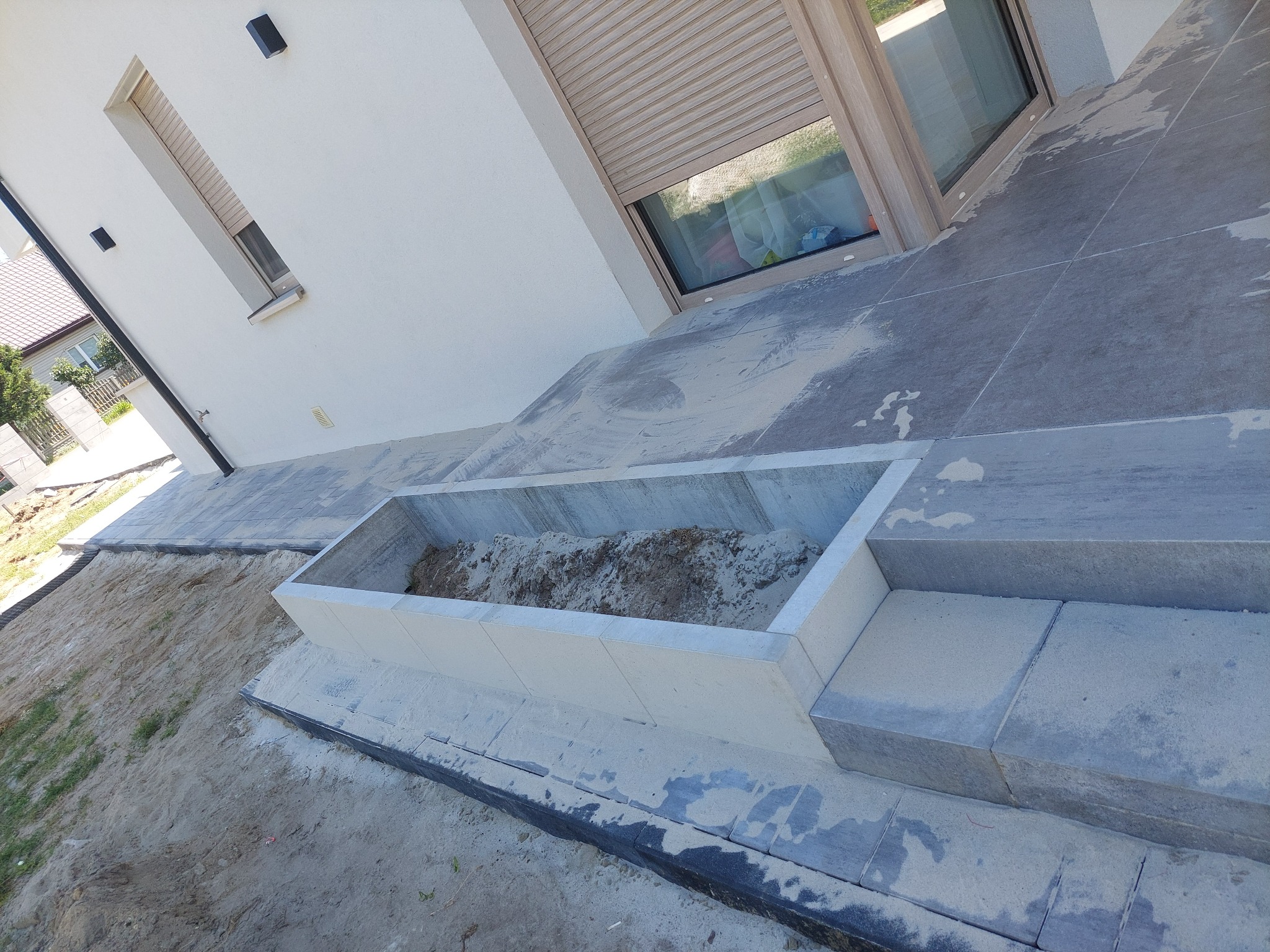 Betonowa donica wbudowana w taras z szarej kostki, wypełniona ziemią, widok z góry na tle białej ściany budynku z oknami z roletami.