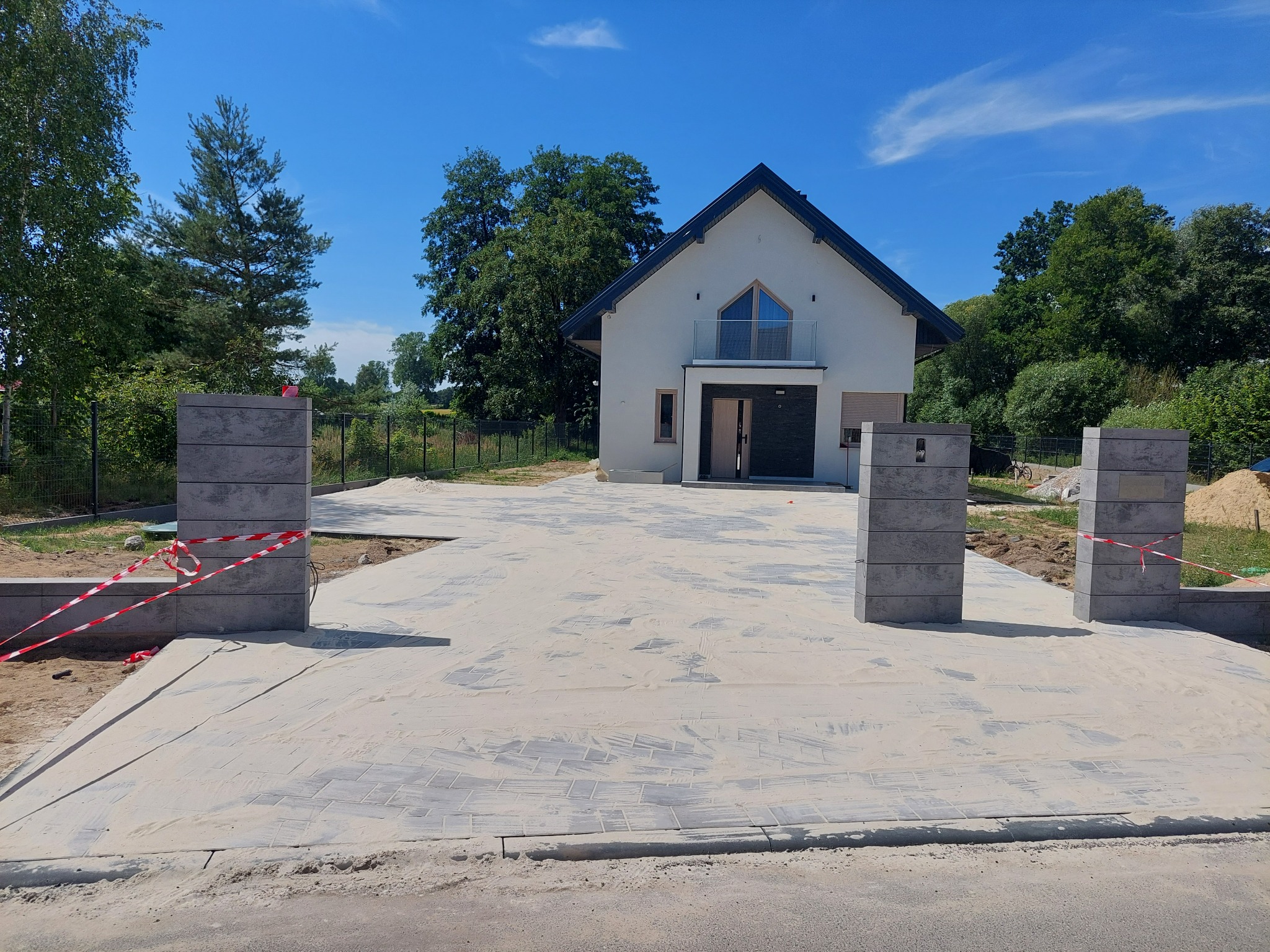Nowoczesny dom jednorodzinny z szarą kostką brukową na podjeździe, widoczne słupki ogrodzeniowe z taśmą ostrzegawczą, błękitne niebo w tle.