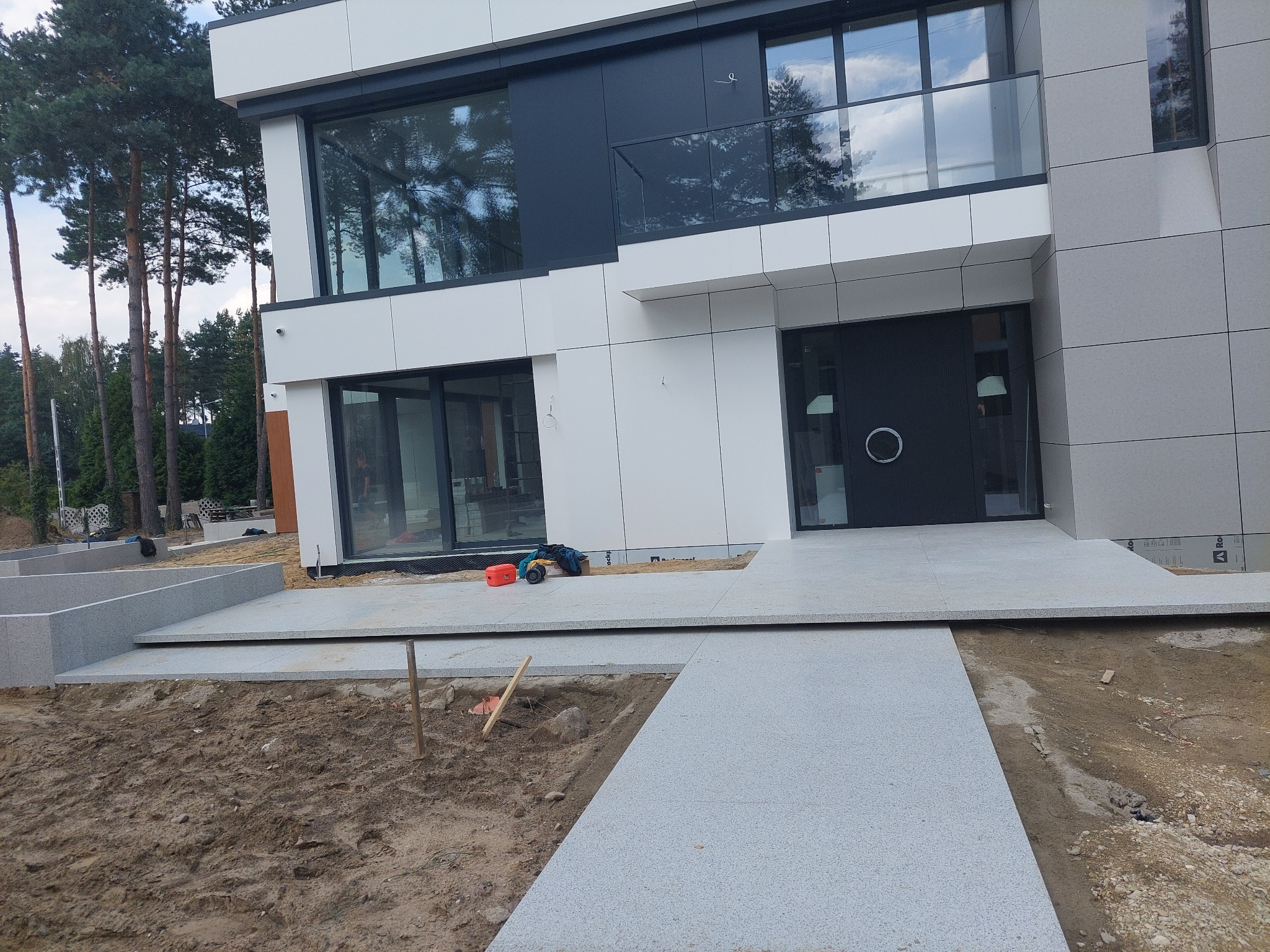 Nowoczesny dom z jasnoszarą elewacją i ścieżką z jasnej kostki granitowej w trakcie budowy, widoczne narzędzia i wykop pod trawnik.