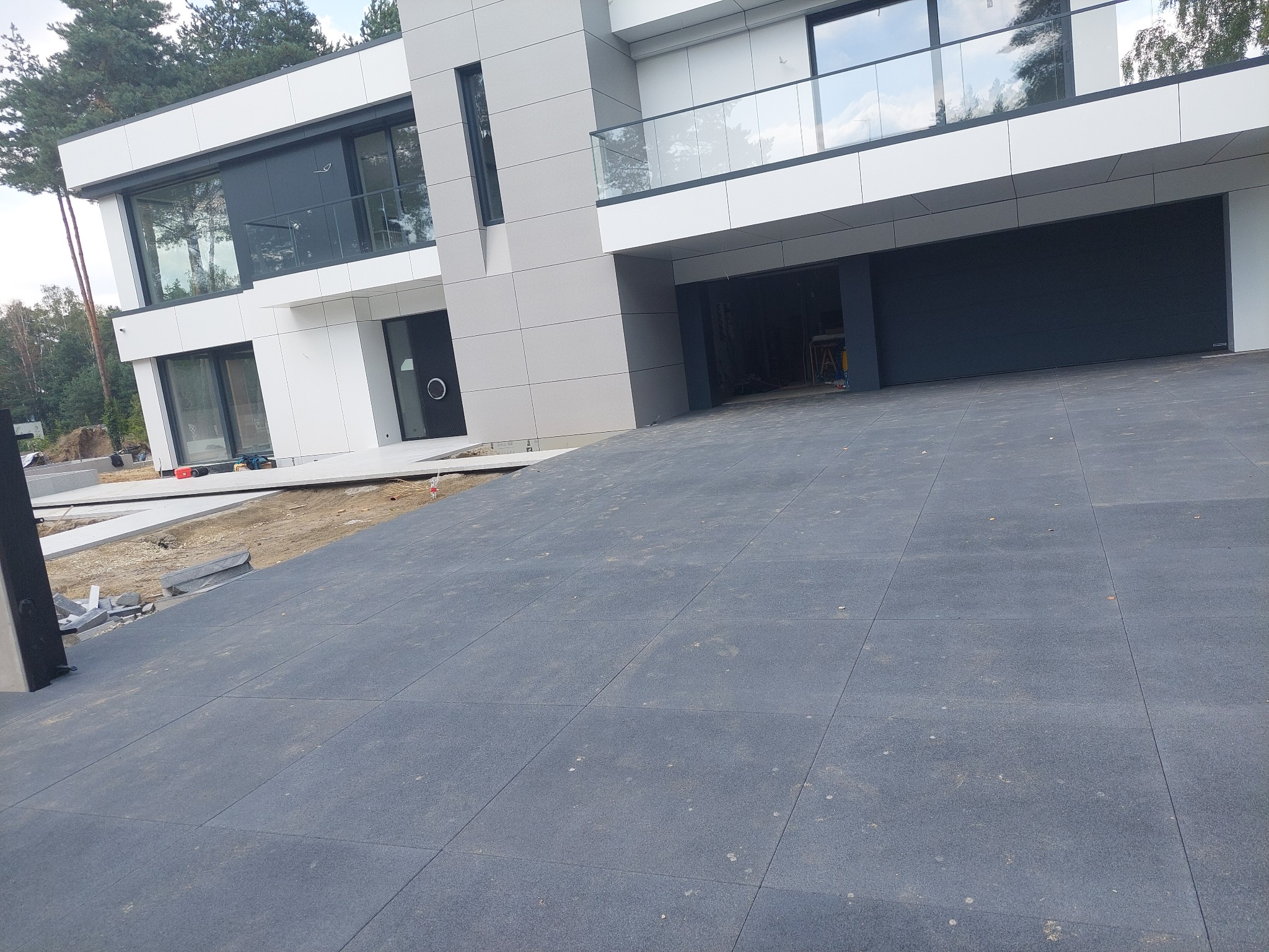 Nowoczesny dom z szarą kostką brukową na podjeździe, widoczne niedokończone prace ziemne obok budynku.