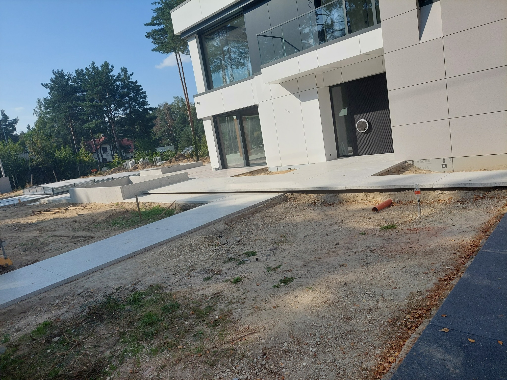 Nowoczesny dom z szarą elewacją i betonowymi ścieżkami w trakcie budowy, widoczne przygotowane podłoże i elementy drenażu z pomarańczową rurą.
