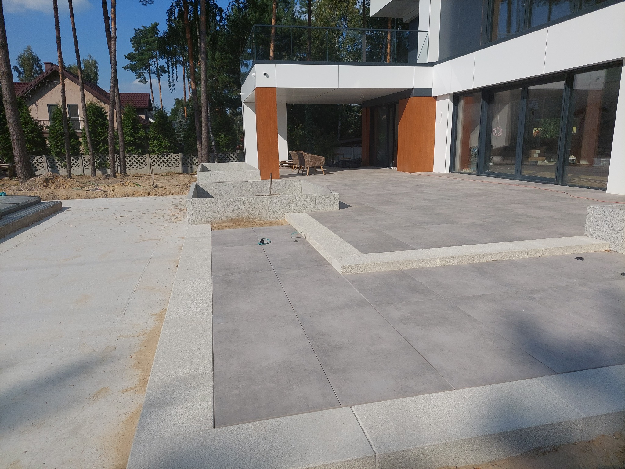 Nowoczesny taras z szarych płyt i jasnych obrzeży, widok na dom z dużymi oknami i zadaszeniem, częściowo w trakcie budowy, widoczne elementy podłoża i ziemi.