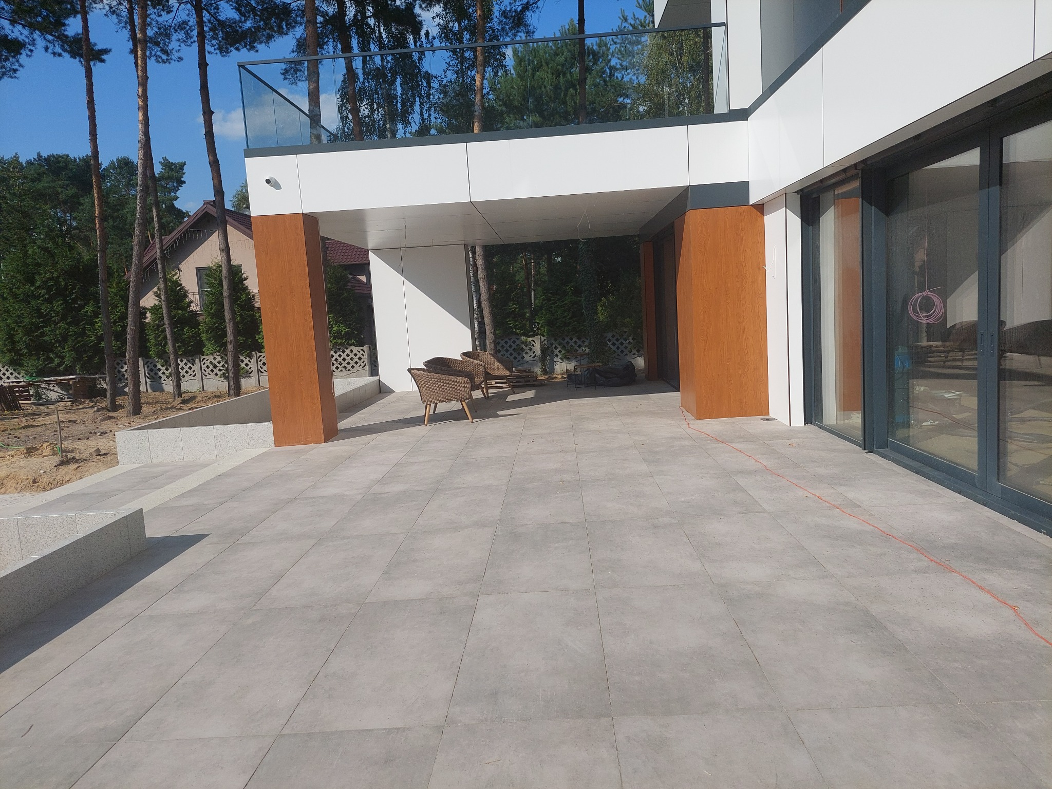 Nowoczesny taras wyłożony szarą kostką, przylegający do budynku z elementami drewna i szkła, z widocznymi meblami ogrodowymi w tle.