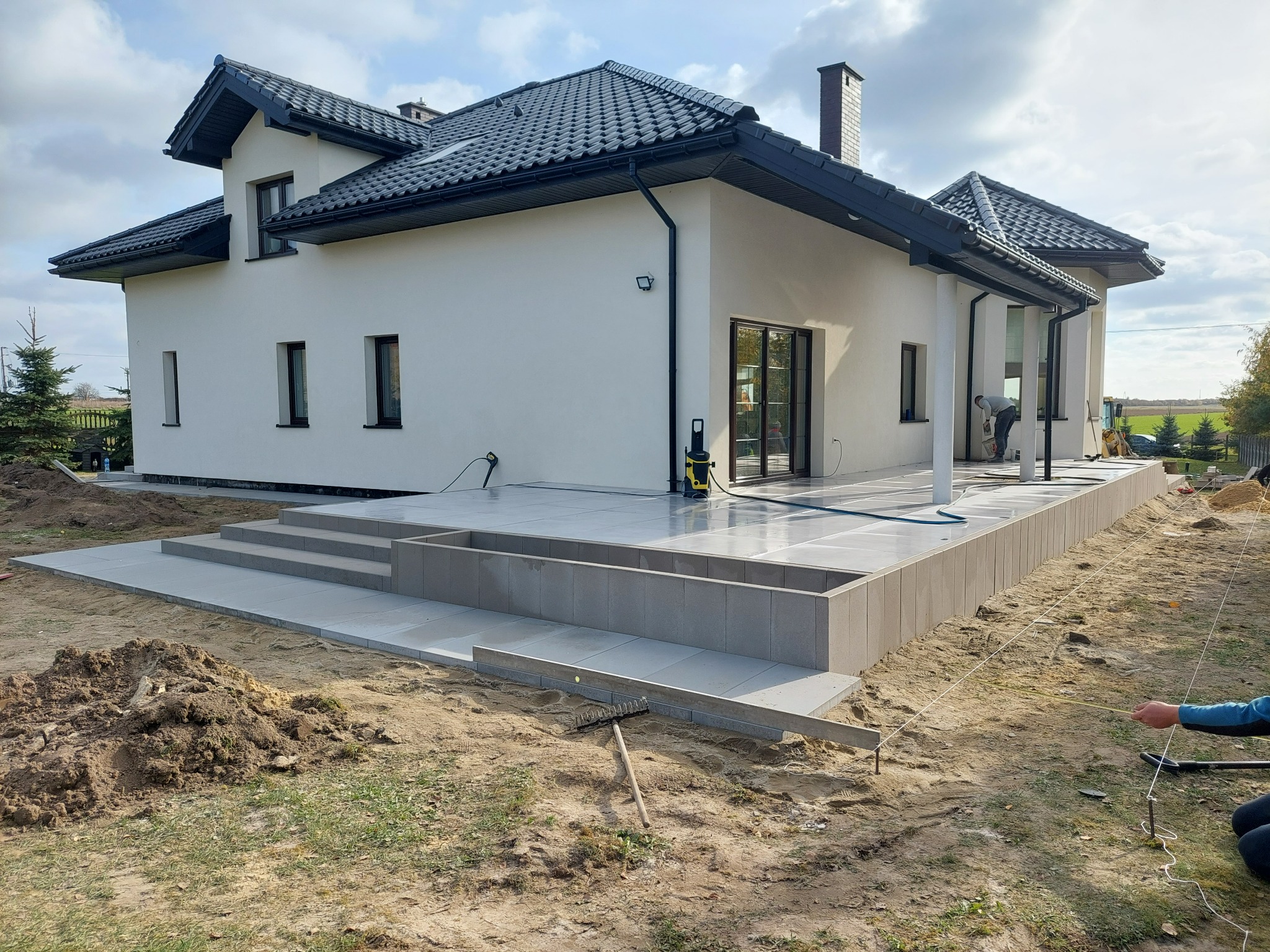 Nowoczesny dom z tarasem w trakcie wykańczania, widoczne narzędzia brukarskie i wytyczone granice, ziemia przygotowana pod dalsze prace.