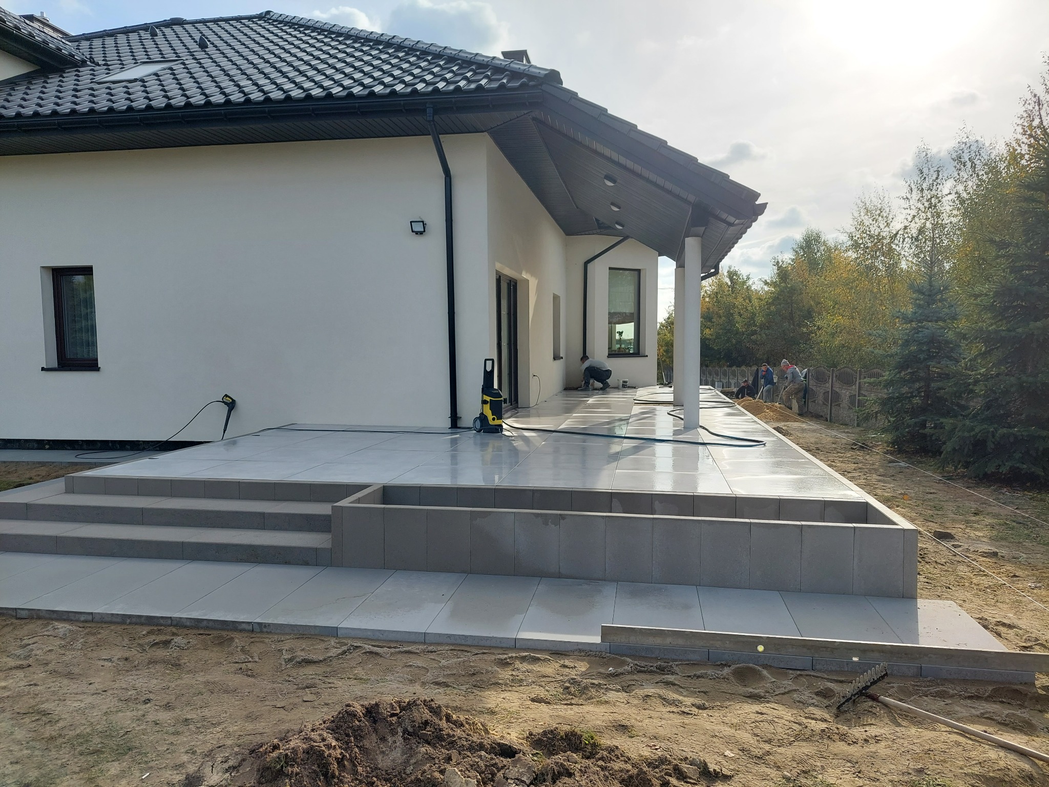 Nowoczesny taras z jasnych płyt, schody z obrzeżami w Woli Moszczenickiej, widoczne prace wykończeniowe i sprzęt do czyszczenia.