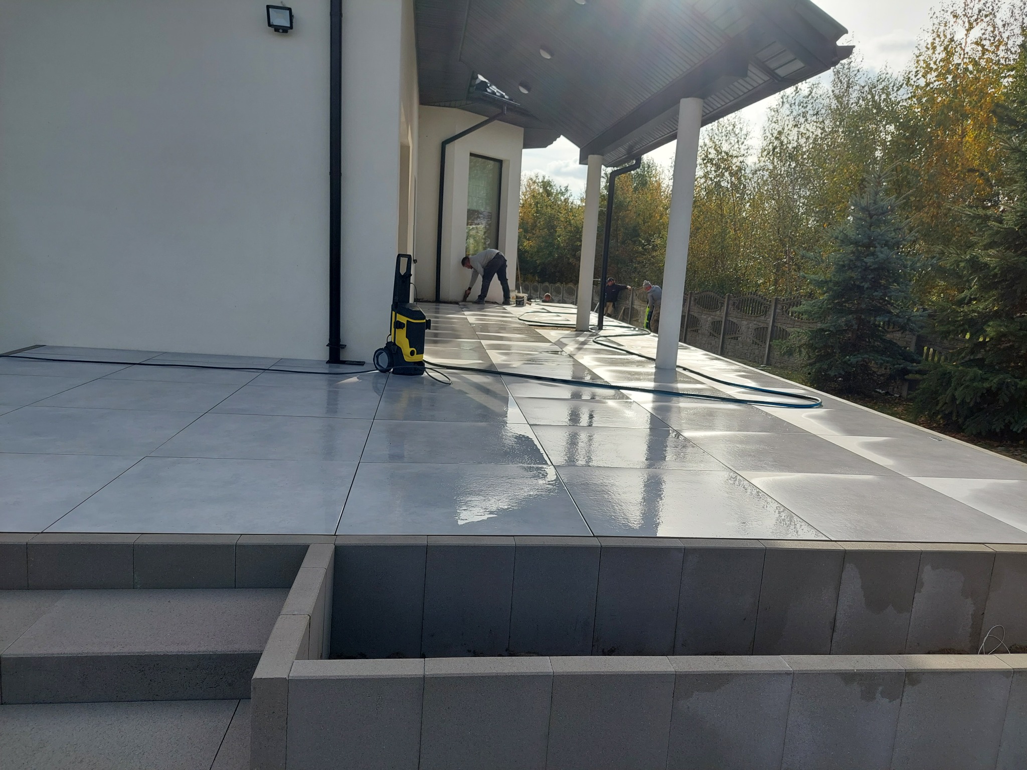 Czyszczenie ciśnieniowe szarej, kwadratowej nawierzchni z płytek na zewnątrz budynku, z widocznymi pracownikami i myjką ciśnieniową w słoneczny dzień.