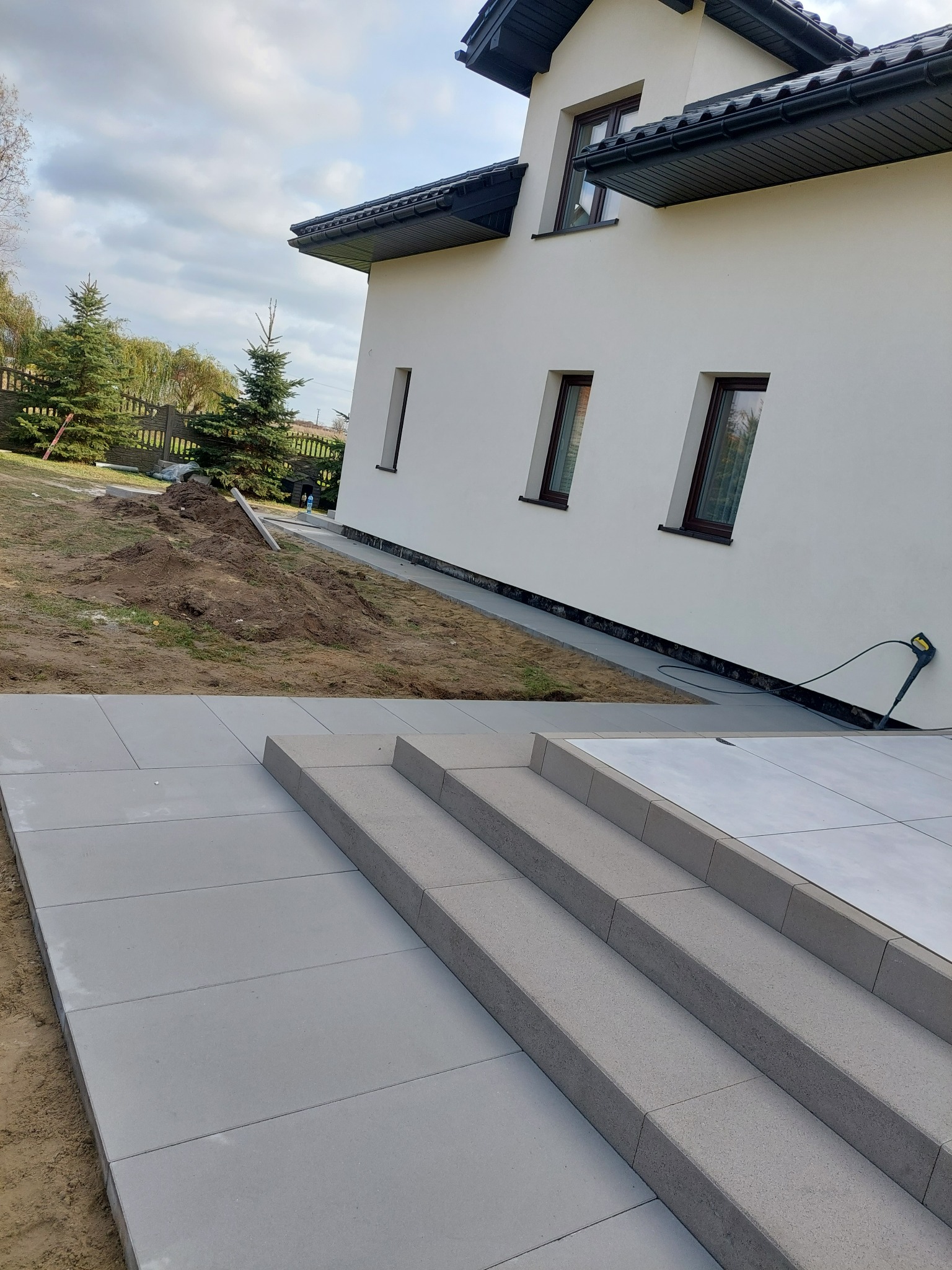 Nowoczesne, szare płytki chodnikowe prowadzące do domu z trzema stopniami, widoczne narzędzie do czyszczenia elewacji przy ścianie.