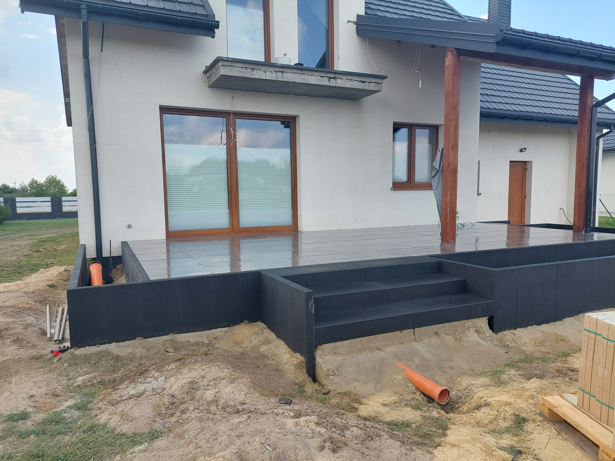 Nowoczesny taras z ciemnej kostki brukowej z schodami, częściowo ukończony, z widocznymi rurami drenażowymi i stertą kostki obok, na tle białego budynku z drewnianą konstrukcją zadaszenia.