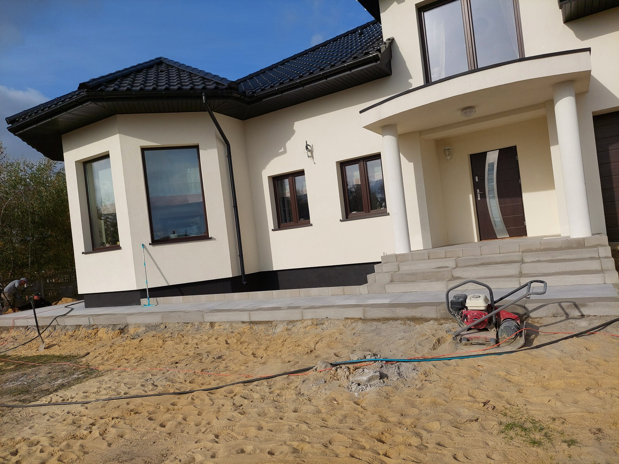 Prace ziemne przy nowym domu: ubijarka spalinowa na piasku, widoczne fundamenty i ułożone obrzeża chodnika z kostki, w tle pracownicy z łopatami.