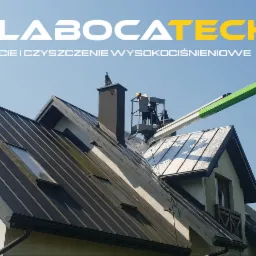 Pracownik w koszu podnośnika czyści dach domu z metalową blachodachówką, widoczne ślady mycia pod ciśnieniem oraz logo firmy na górze kadru.