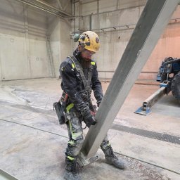 LT Labocatech Usługi wysokościowe, usługi alpinistyczne - Pracownik w kasku i uprzęży, w trakcie prac wysokościowych wewnątrz hali, trzymający metalowy element konstrukcyjny. W tle widoczny podnośnik i drabina.