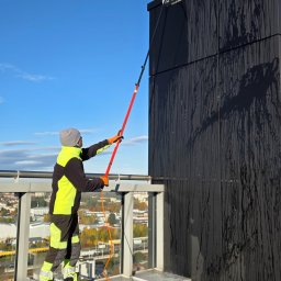 LT Labocatech Usługi wysokościowe, usługi alpinistyczne - Pracownik w odzieży ochronnej myje ciemną elewację budynku za pomocą szczotki na długim wysięgniku. Widoczne zacieki i panorama miasta w tle.