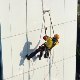 LT Labocatech Usługi wysokościowe, usługi alpinistyczne - Pracownik w kasku, na linach, myje białą elewację budynku za pomocą specjalistycznego sprzętu. Widok z góry, słoneczny dzień.